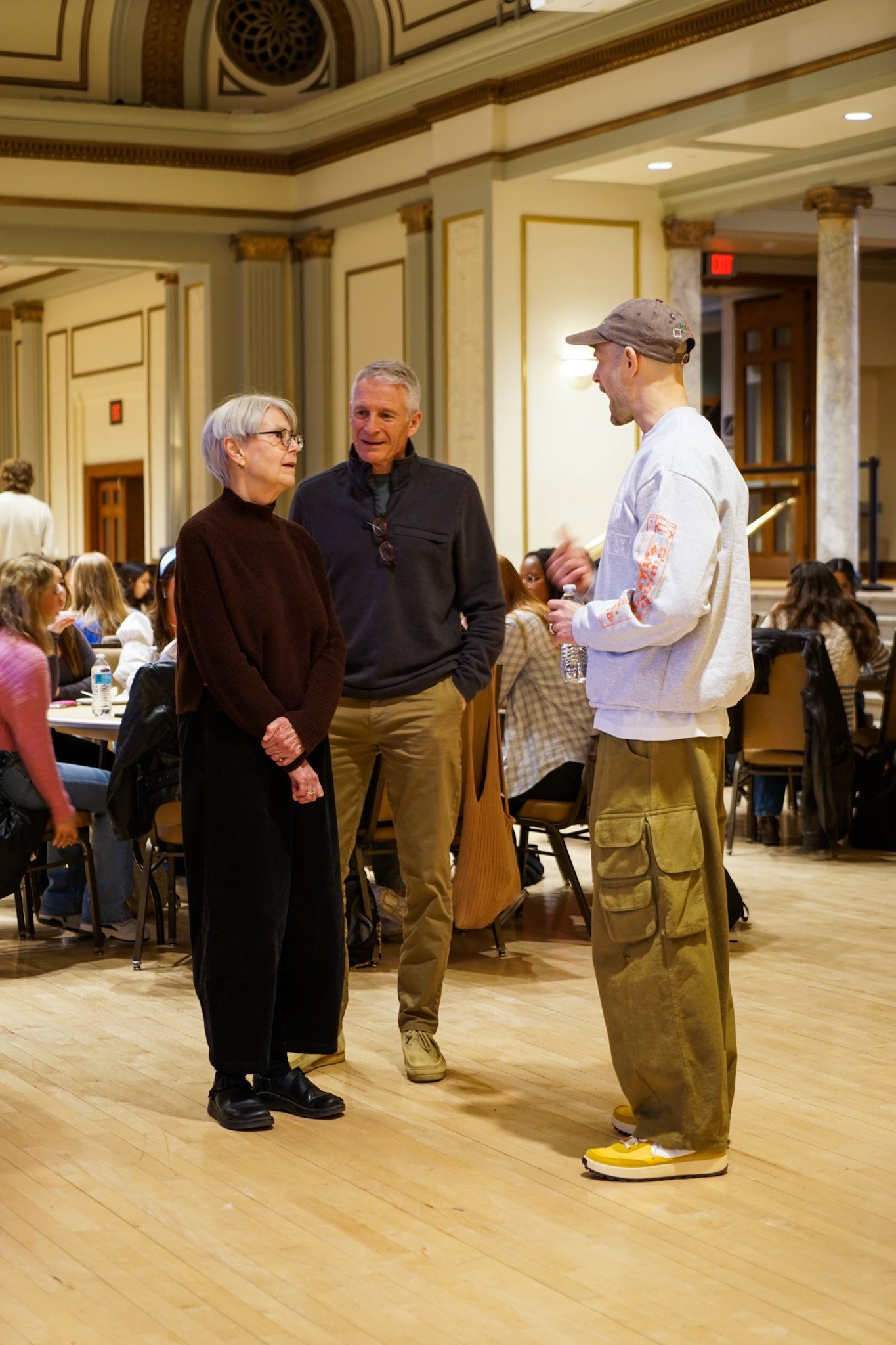 Three people engaged in conversation inside a large, elegant room filled with seated attendees at tables.