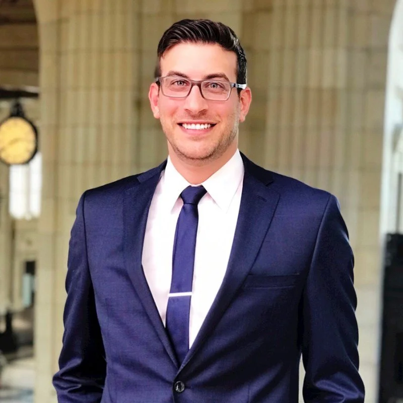 A young man in a navy suit, white shirt, and navy tie, smiling at the camera indoors with columns and a clock in the background.
