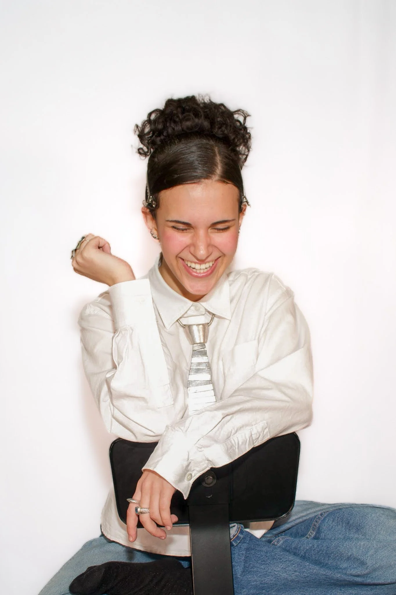 A young woman with curly hair and a hair clip, wearing a white shirt and a striped tie, sitting with her arms crossed and laughing, against a white background.