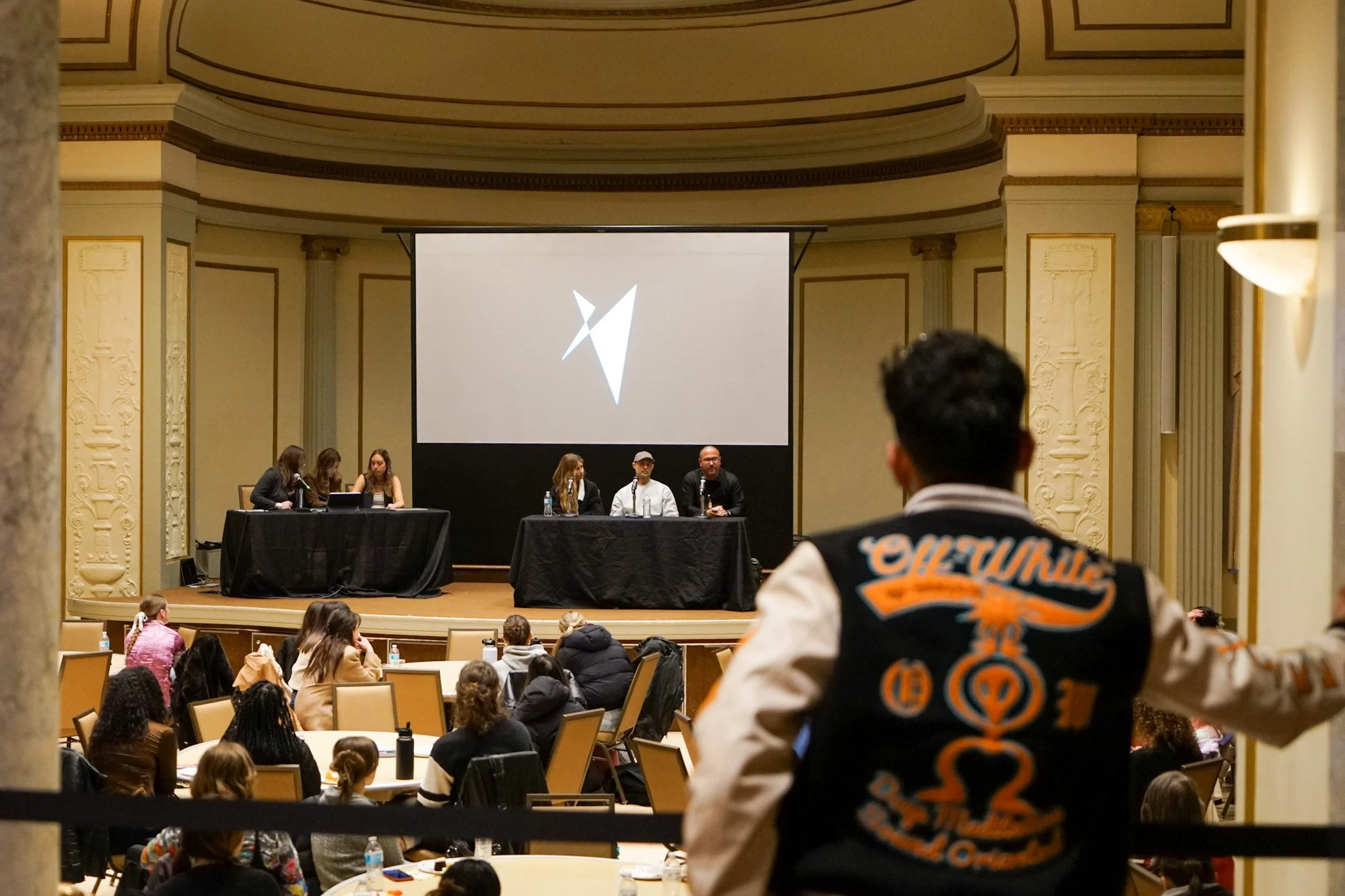 A panel of five people on stage at a conference or panel discussion, with an audience seated in front, and a person in the foreground wearing a varsity jacket.