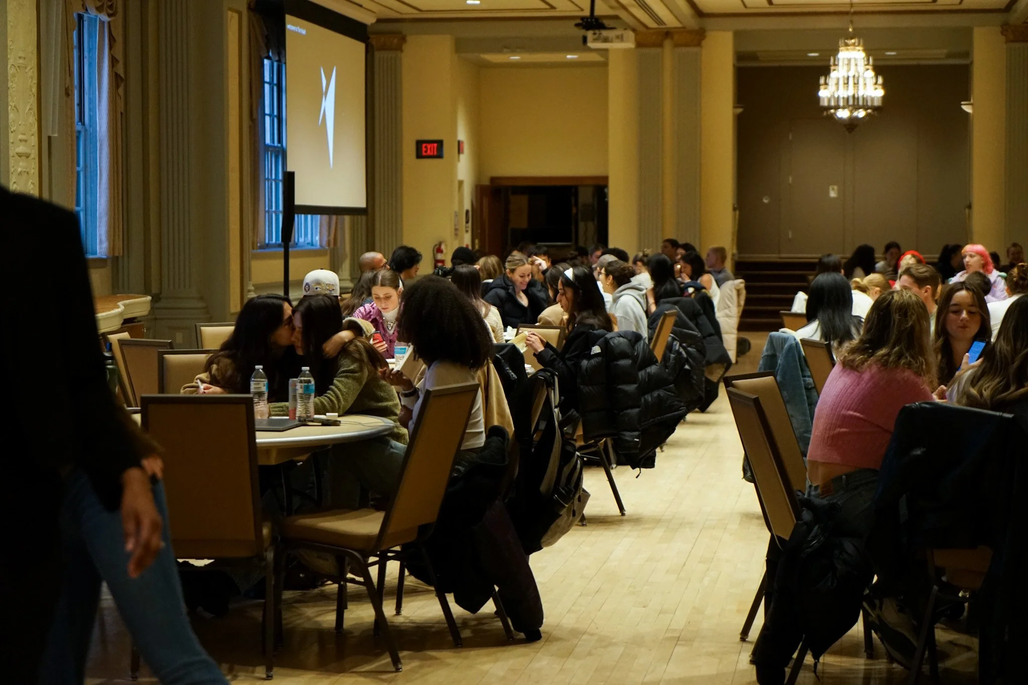 A large conference room filled with people sitting at round tables, some engaged in conversation and others using their phones, with a stage area at the front and large windows with curtains on the side.