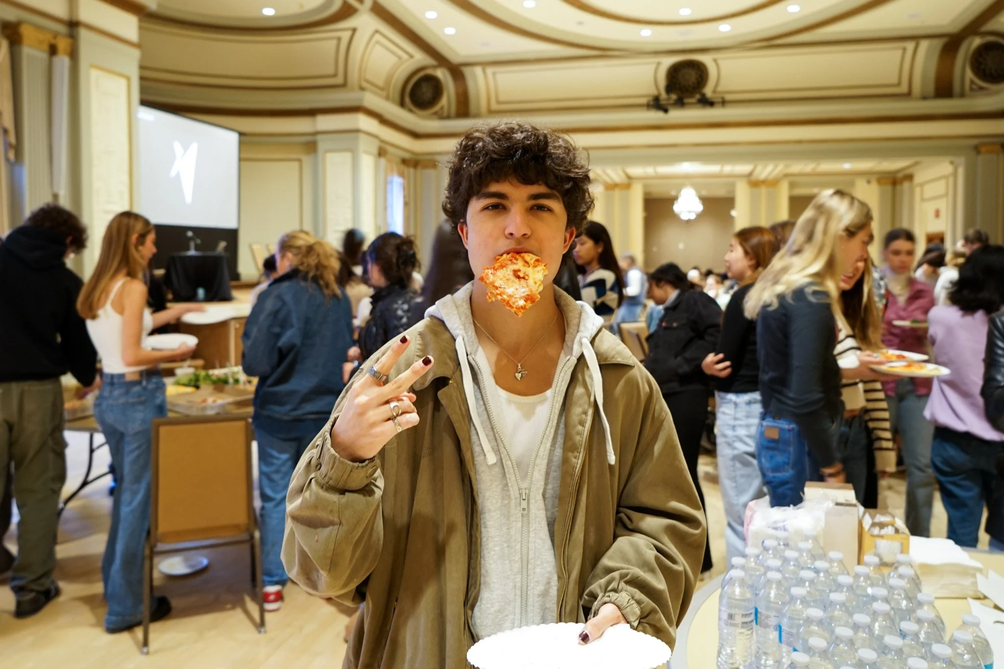 A young person with curly dark hair, wearing a tan jacket and white hoodie, posing with two fingers up, holding a slice of pizza in front of their mouth in an indoor setting with many people around, some serving food at a buffet table.