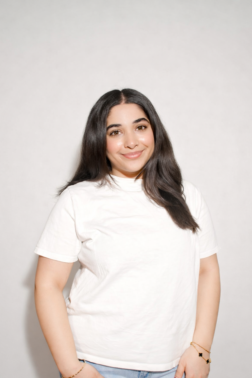 A young woman with long dark hair smiling, wearing a white t-shirt and jewelry, standing against a light-colored wall.