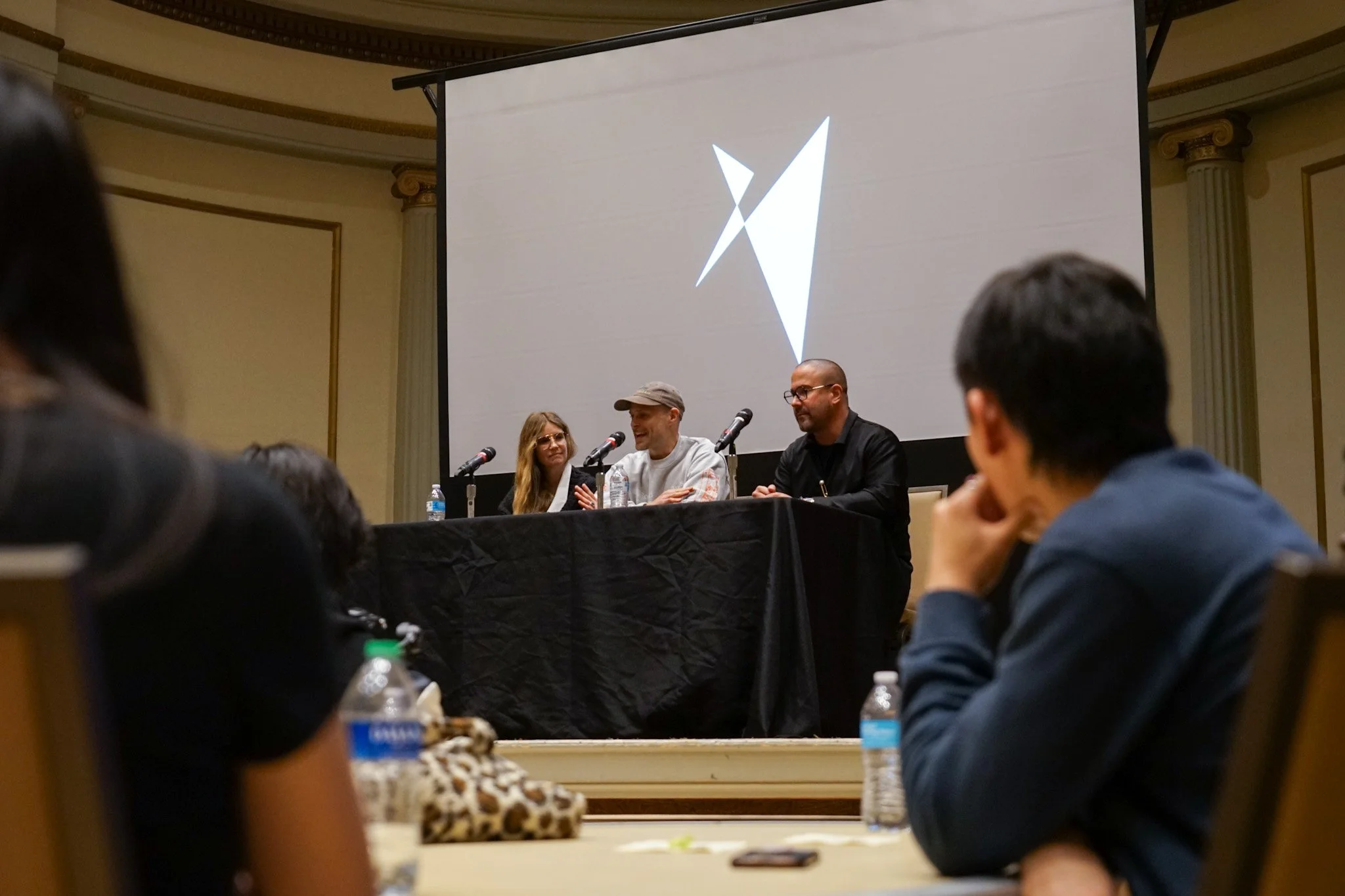 A panel of three people seated behind a table on a stage at a conference, with a large projection screen behind them displaying a stylized abstract logo. Several audience members are visible in the foreground.