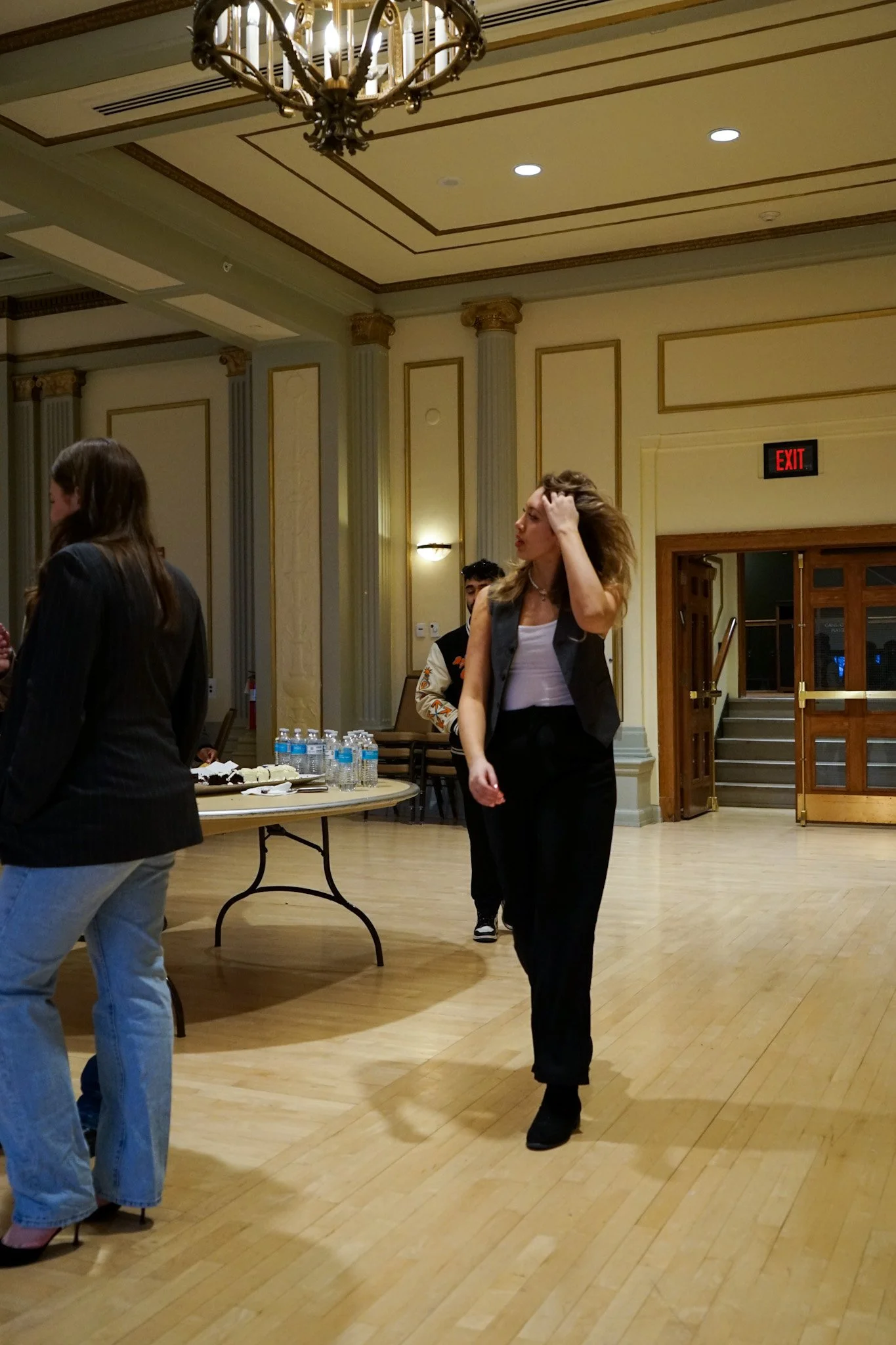 Interior of a formal event room with ornate moldings and lighting, people standing and walking around, including a woman with long hair touching her head, near a table with water bottles and snacks.