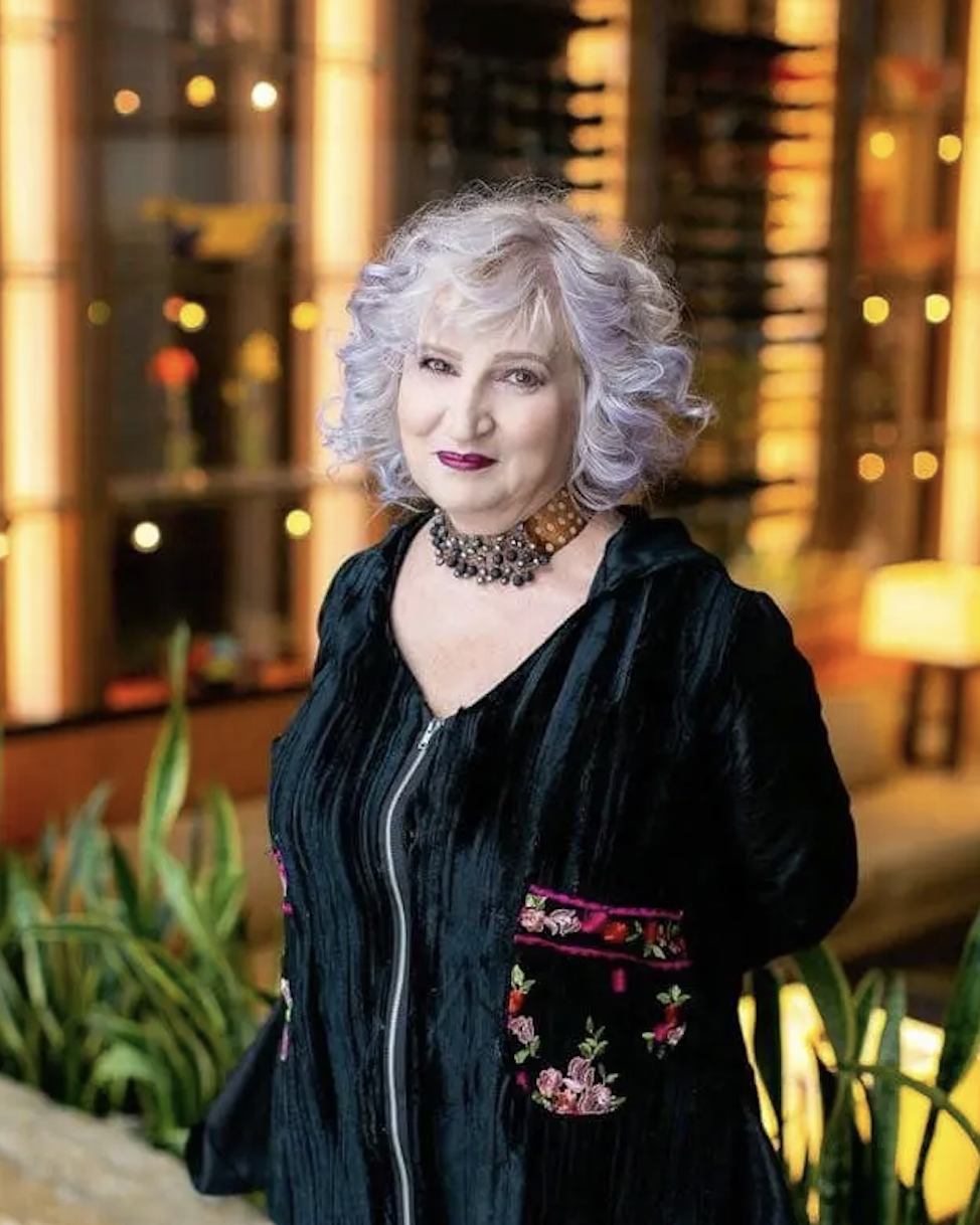 An older woman with curly, silver hair in a black velvet dress with embroidered floral patches, wearing a beaded choker necklace, standing indoors in front of a background with warm lighting and bookcases.