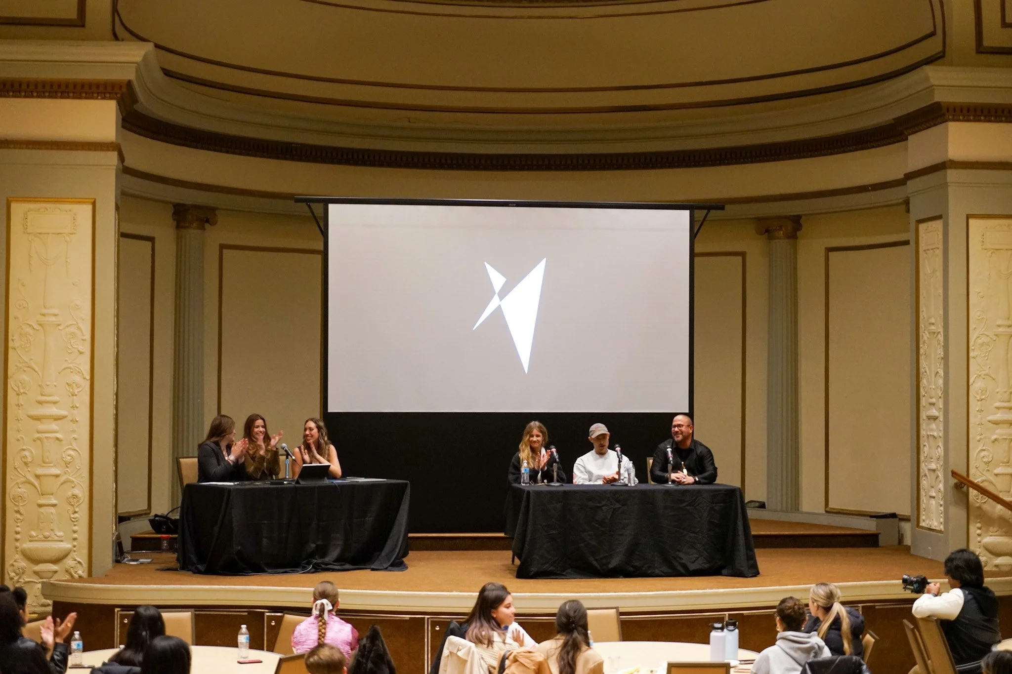Panel discussion on a stage in a grand hall with an audience, featuring five people at two tables with microphones, and a large screen behind displaying a stylized geometric logo.