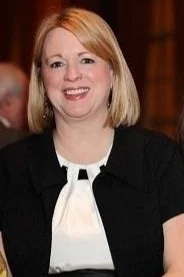 A woman with shoulder-length blonde hair wearing a black top with a white detail, smiling in an indoor setting.