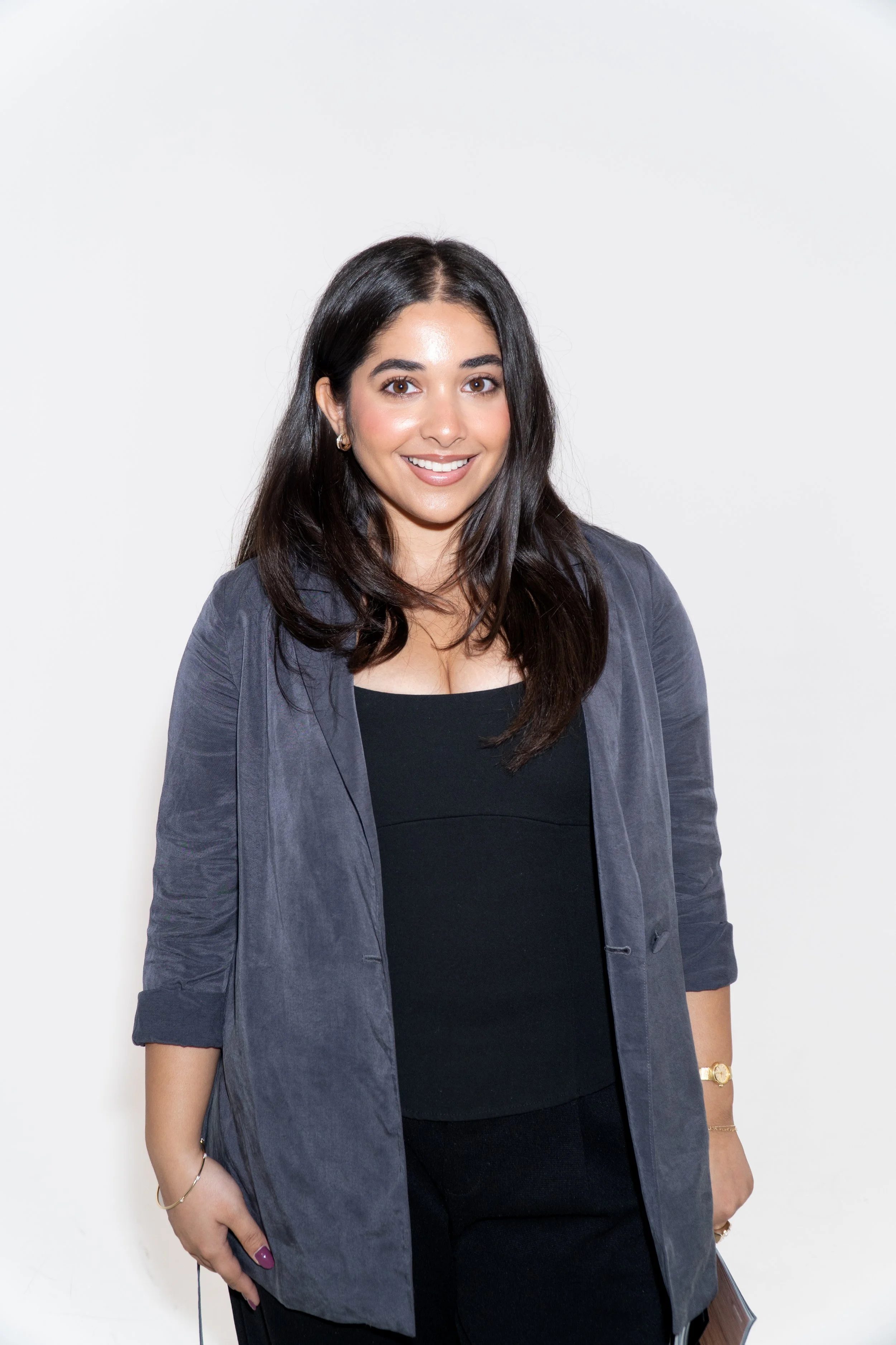 A young woman with long dark hair smiling, wearing a white t-shirt and jewelry, standing against a light-colored wall.