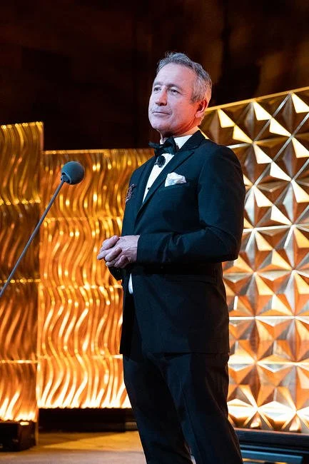 Man in a tuxedo standing at a microphone on stage with a gold geometric backdrop.