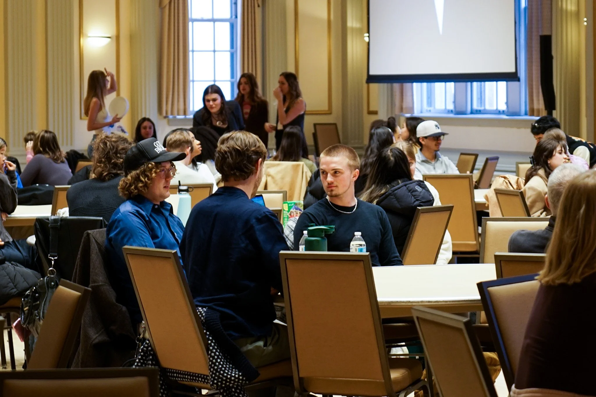 A large group of young people sitting and talking in a conference or event room with high windows, beige walls, and a projection screen.