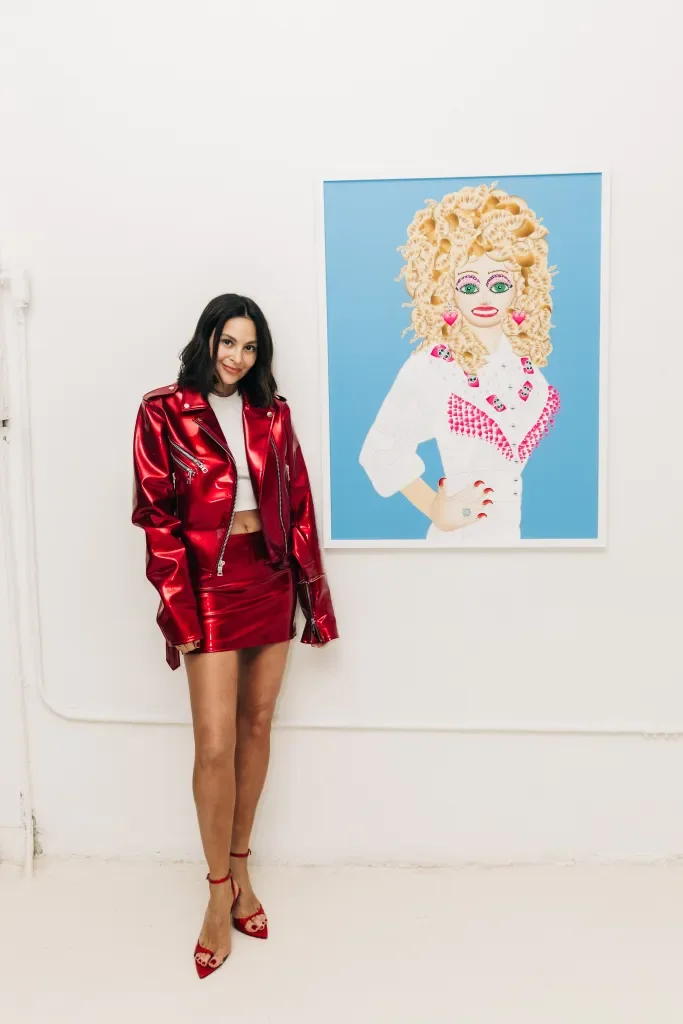 Young woman holding a black hat and bag, wearing a long, striped shirt and jeans, standing on a city sidewalk near a brick building.