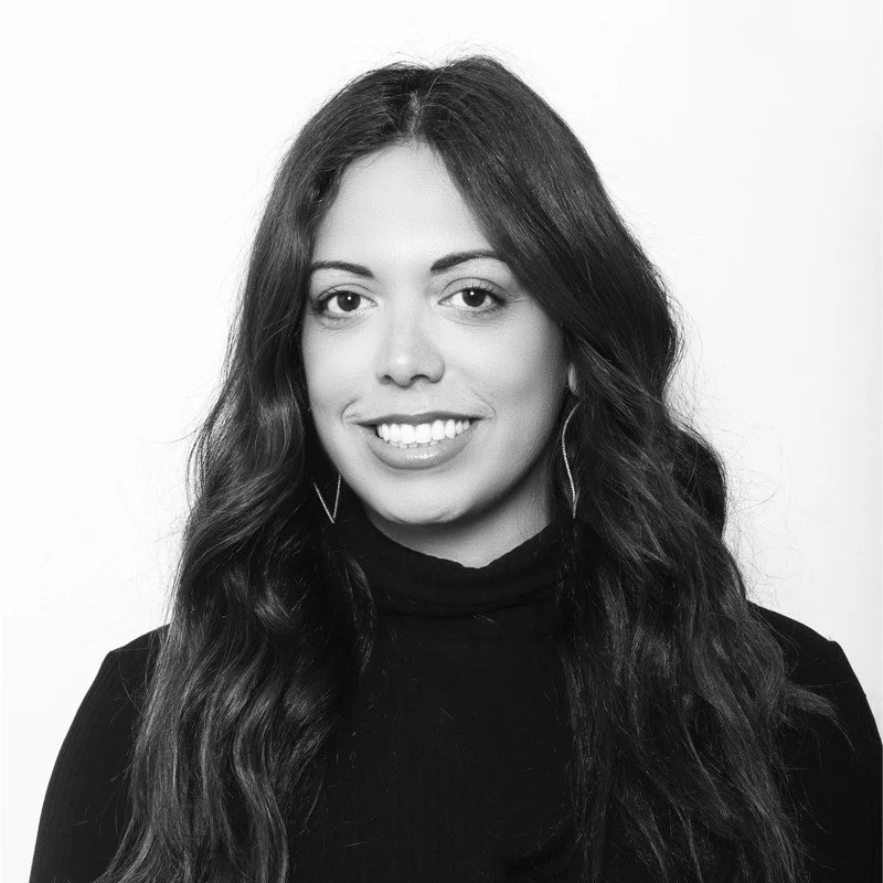 Black and white portrait of a young woman with wavy hair, wearing a black top and earrings, smiling at the camera.