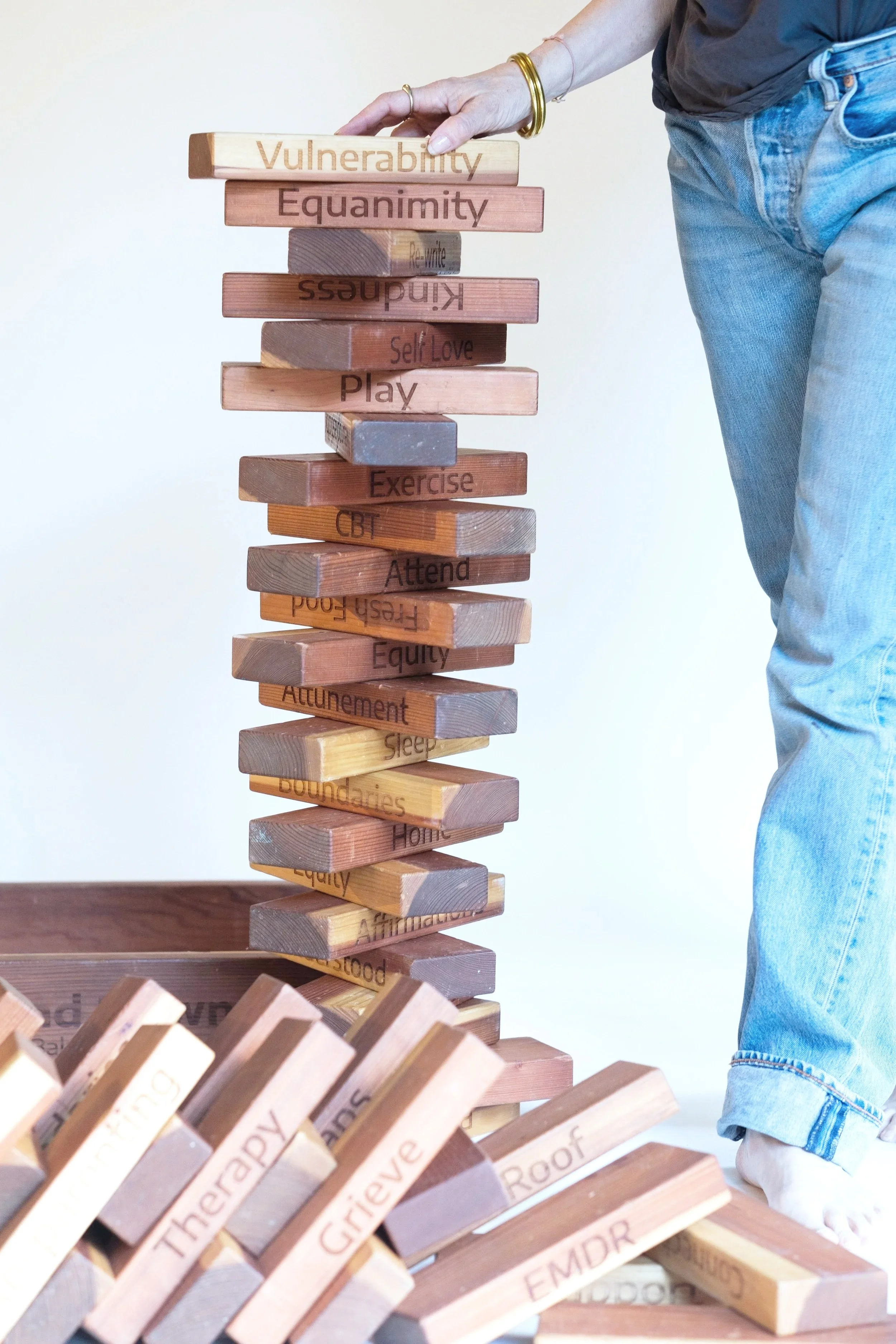 A person is stacking wooden blocks with words related to mental health and wellness, including 'Vulnerability,' 'Equaninity,' 'Self Love,' and 'Play.' Some blocks are fallen on the floor, and the person is wearing blue jeans and a dark shirt.