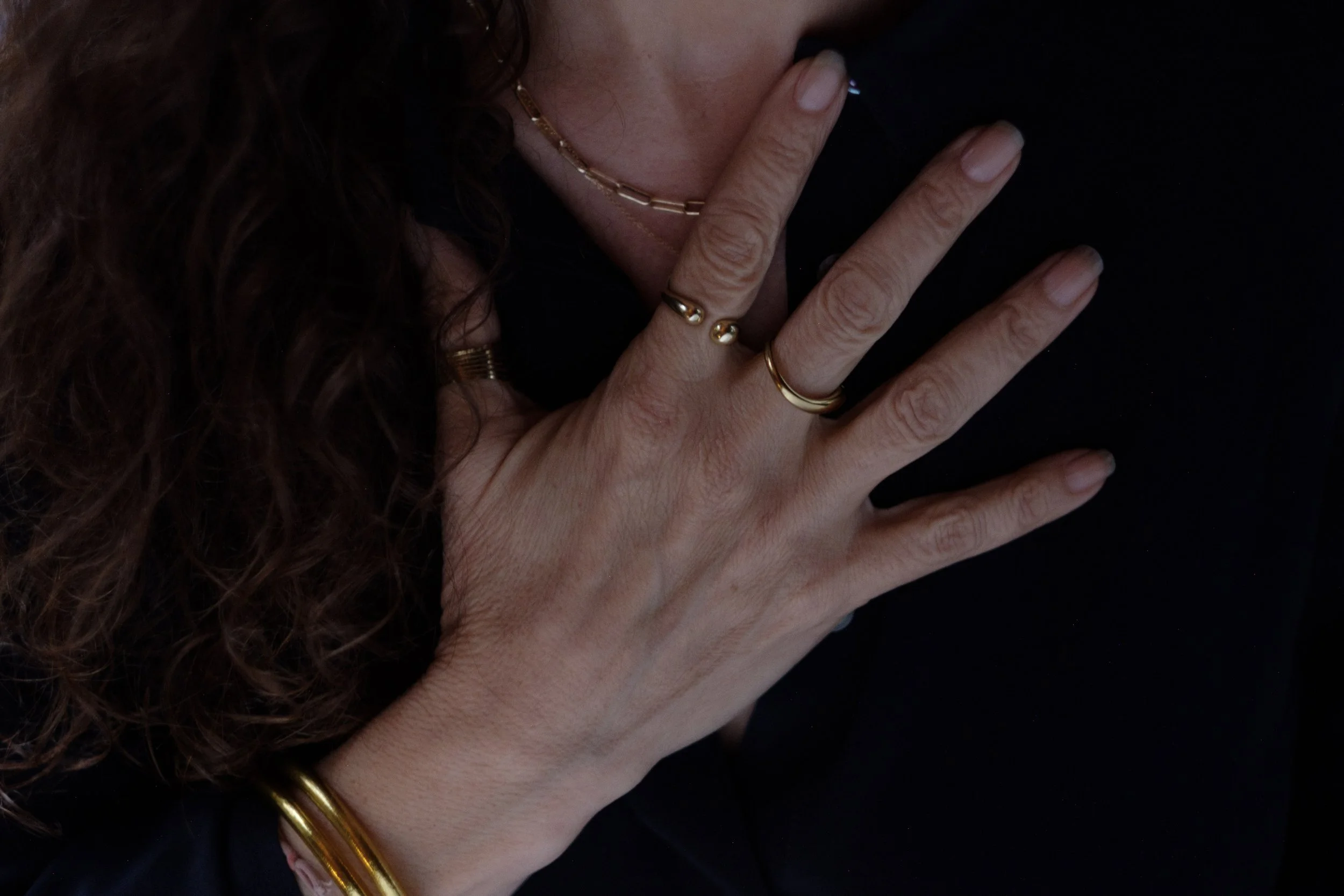 Close-up of a woman with curly hair wearing jewelry, including rings, a necklace, and a bracelet, with her hand near her face and her mouth touching her finger against a black background.
