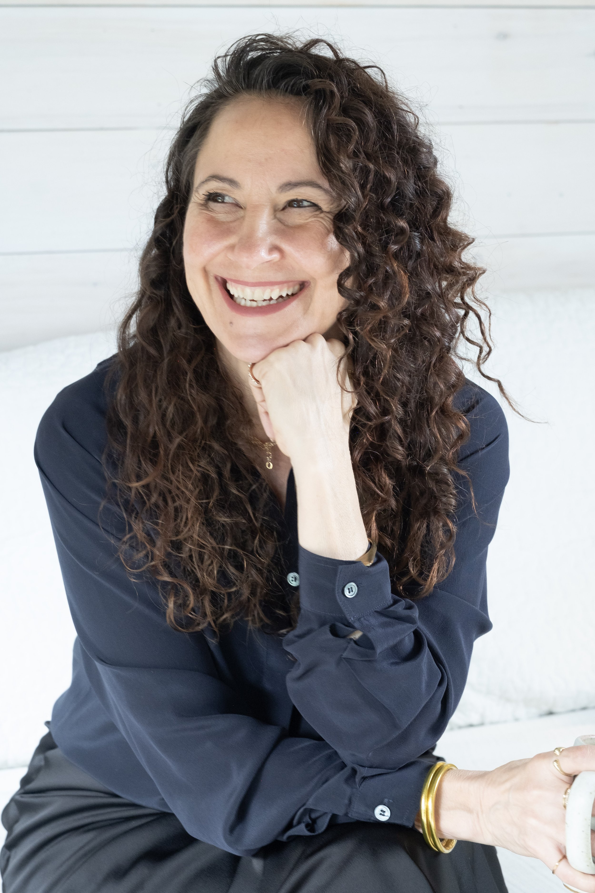 A woman with curly brown hair smiling brightly, wearing a navy blue blouse and gold jewelry, sitting against a white wall and looking to her right.