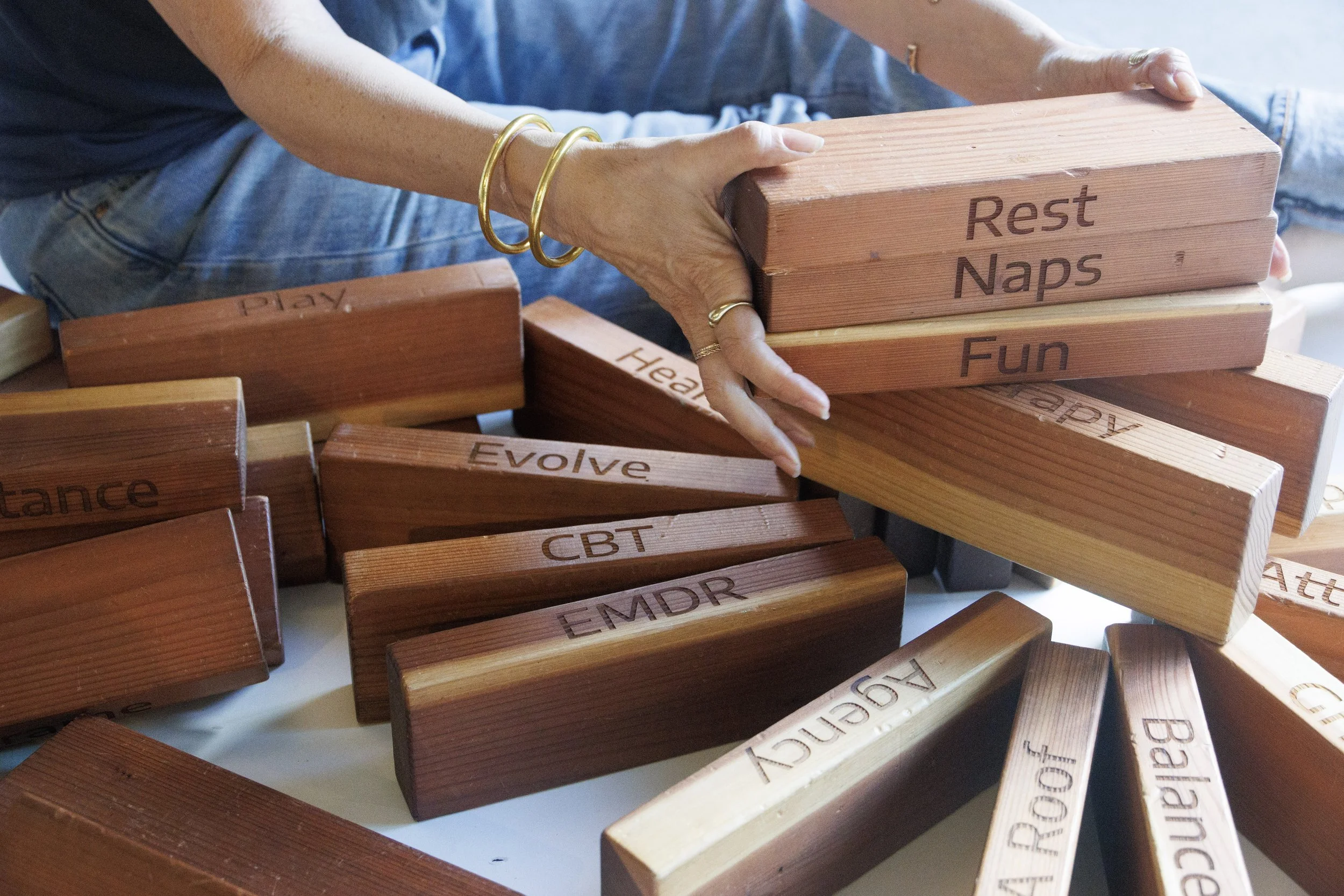 A person stacking wooden blocks with positive affirmations like 'Rest Naps,' 'Fun,' and 'Play,' on a table.