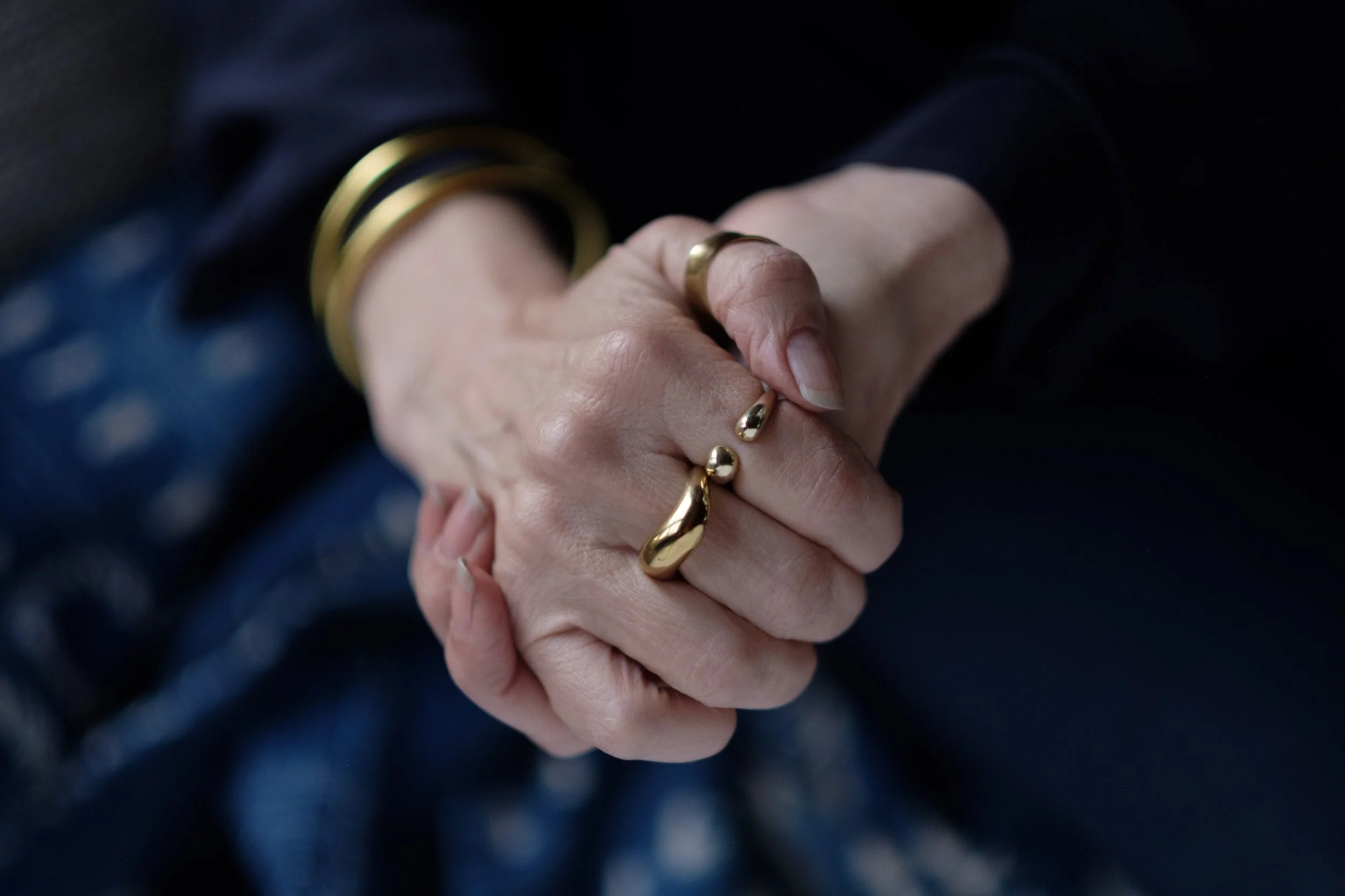 Close-up of two hands clasped together, wearing multiple gold rings and gold bracelets.
