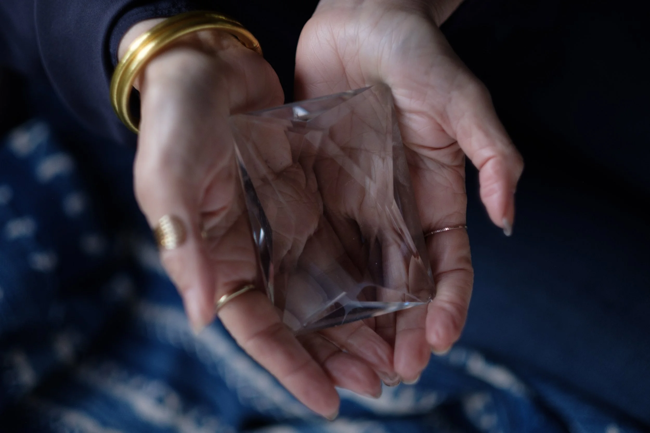Close-up of a hand holding a piece of clear, faceted glass or crystal, with multiple angles and reflections, against a dark background.