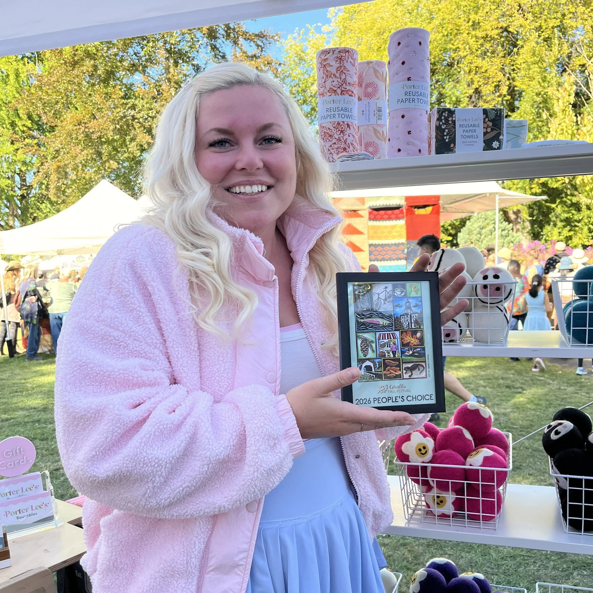 A woman smiling. The woman is holding a framed Corvallis Fall Festival poster that reads "2026 People's Choice."