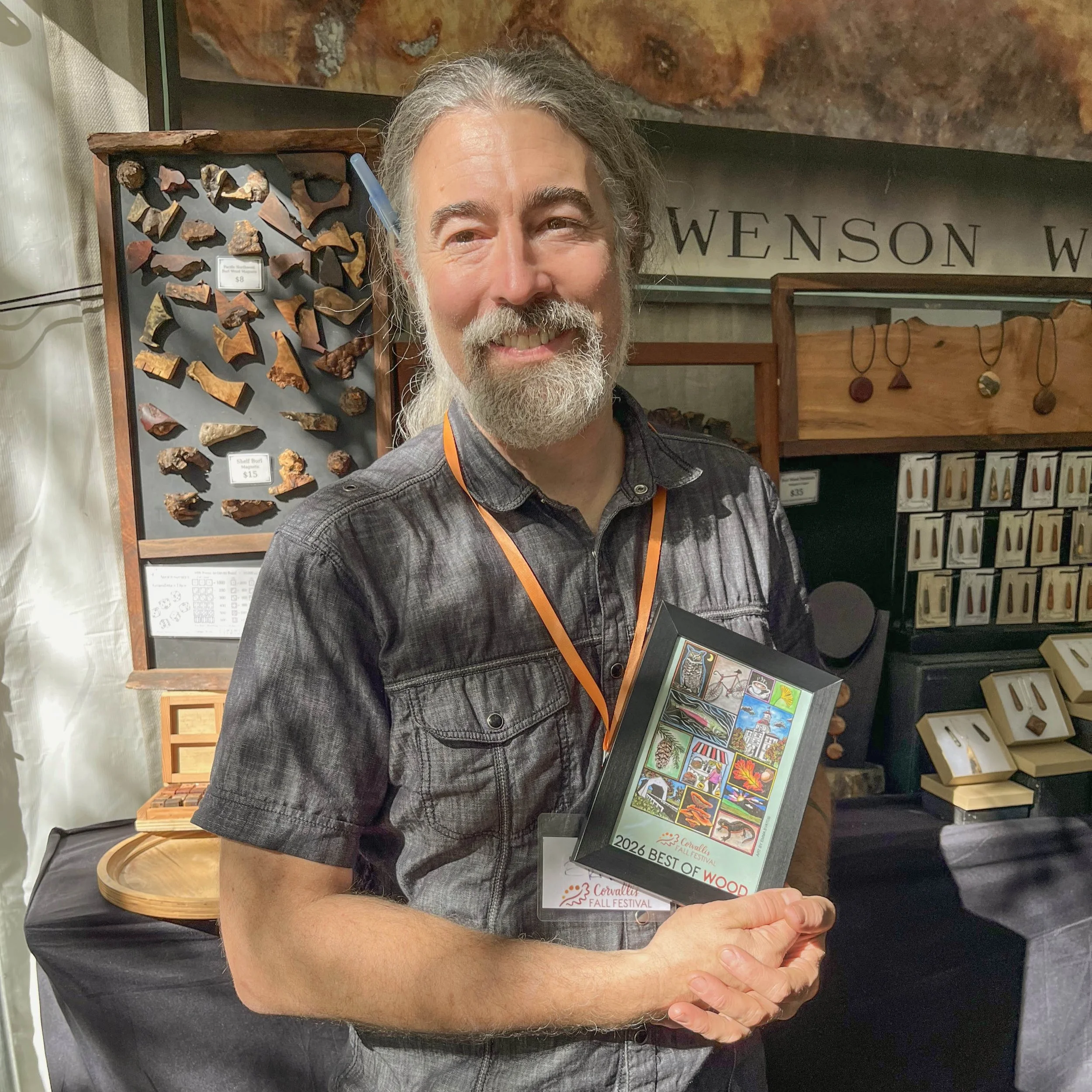 A man smiling and holding a framed Corvallis Fall Festival poster that reads "2026 best of wood."