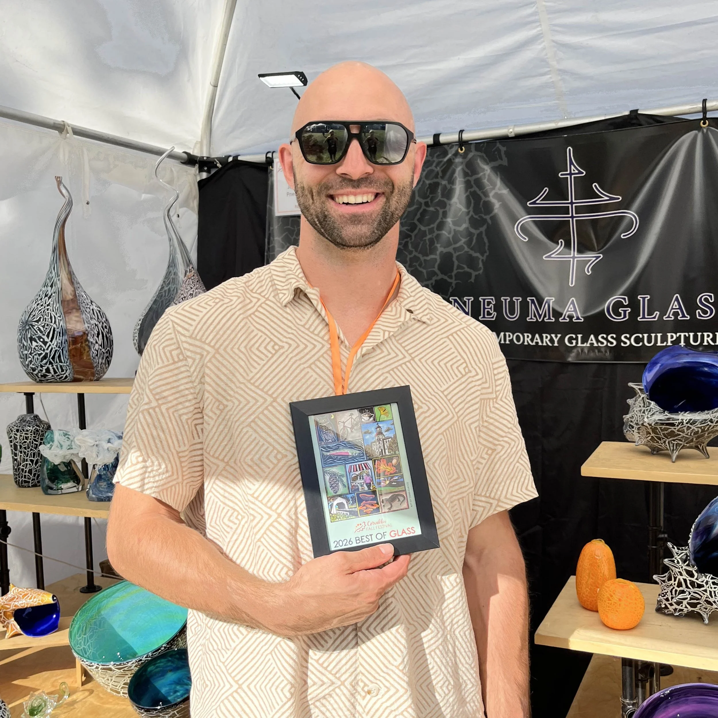 A man smiling and holding a framed Corvallis Fall Festival poster that reads "2026 Best of Glass."