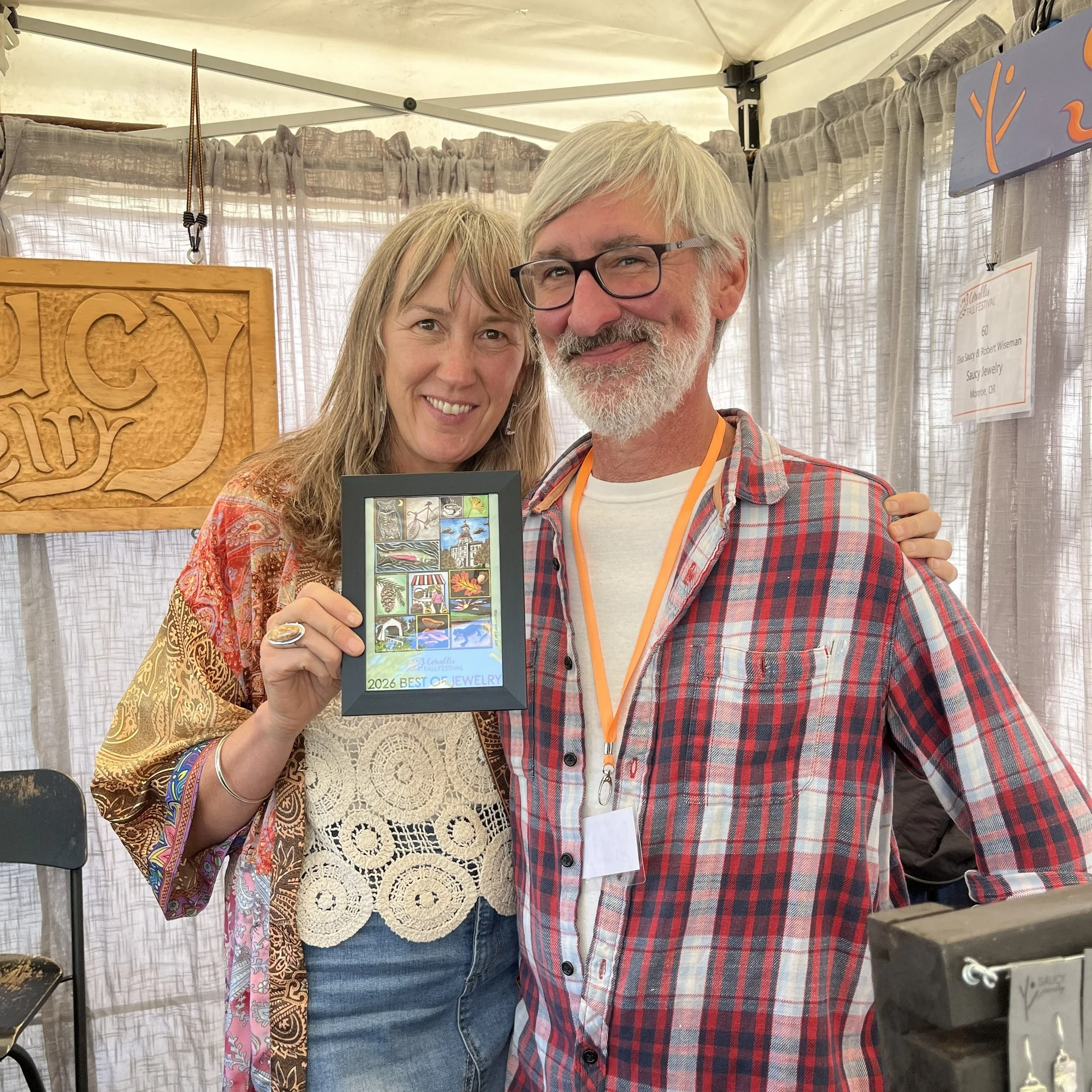A man and women smiling and holding a framed Corvallis Fall Festival poster that reads "2026 best of jewelry."