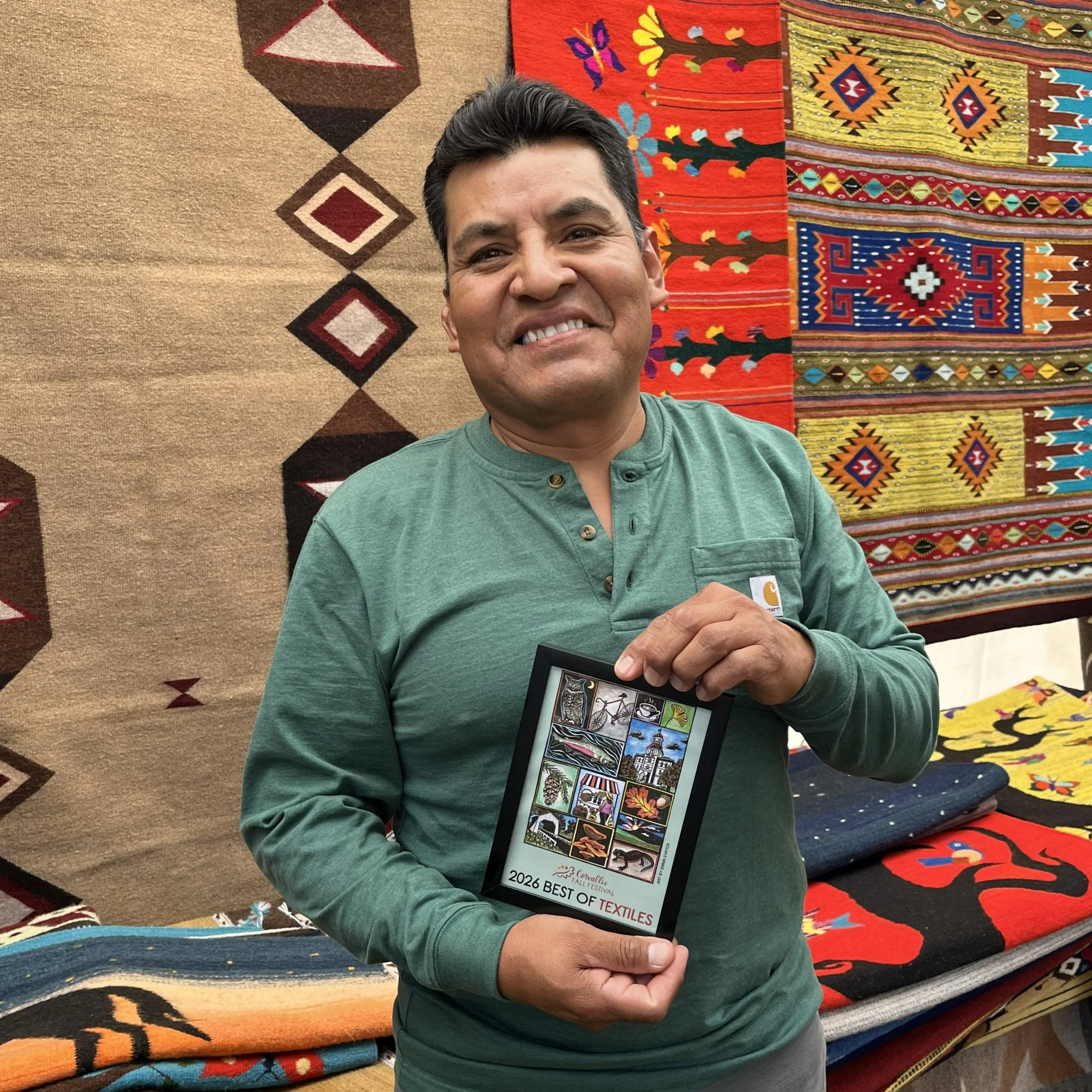 A man smiling in front of colorful textile art and holding a framed Corvallis Fall Festival poster that reads "2026 best of textiles."