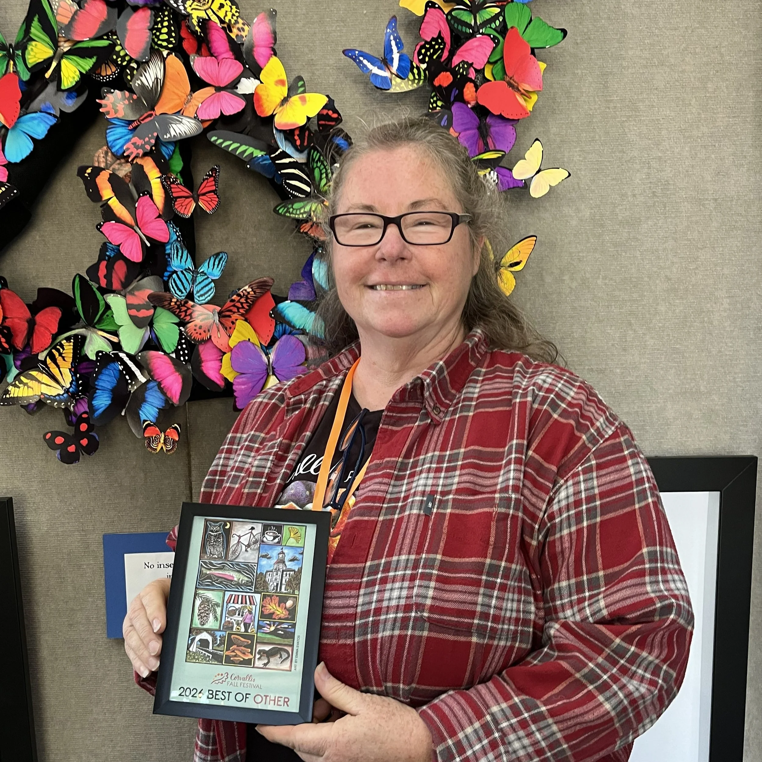 A woman smiling and holding a framed Corvallis Fall Festival poster that reads "2026 best of other"