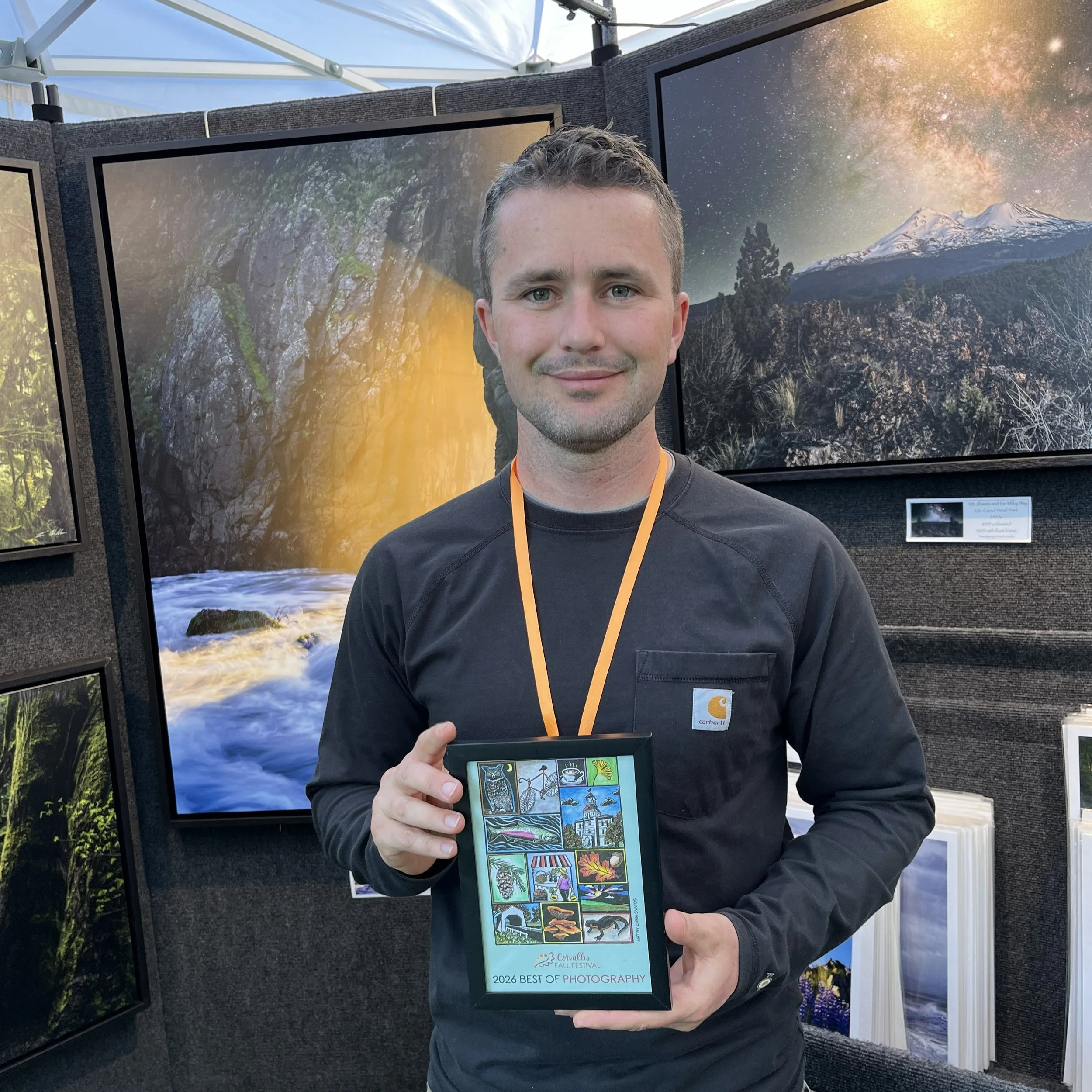 A man holding a framed Corvallis Fall Festival poster that reads "2026 best of photography."
