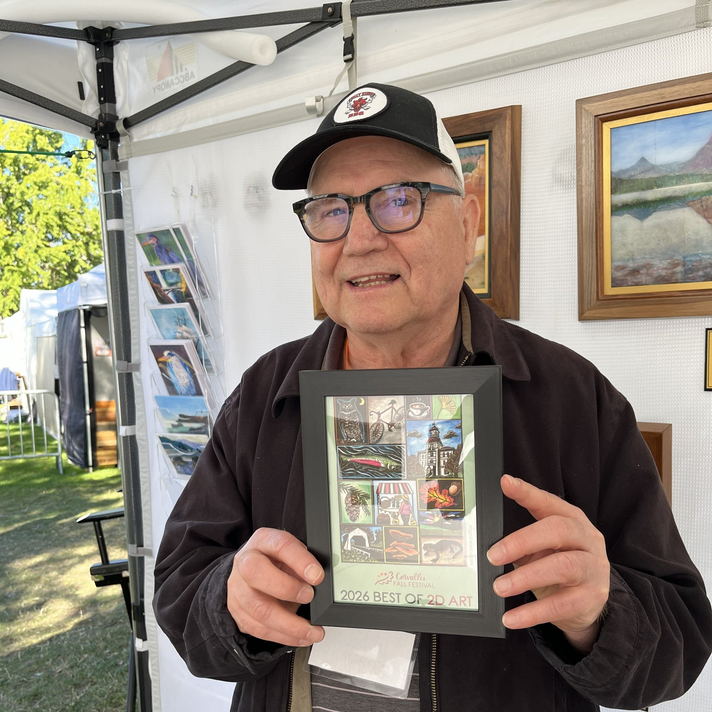 A man smiling and holding a framed Corvallis Fall Festival poster that reads "2026 best of 2D."