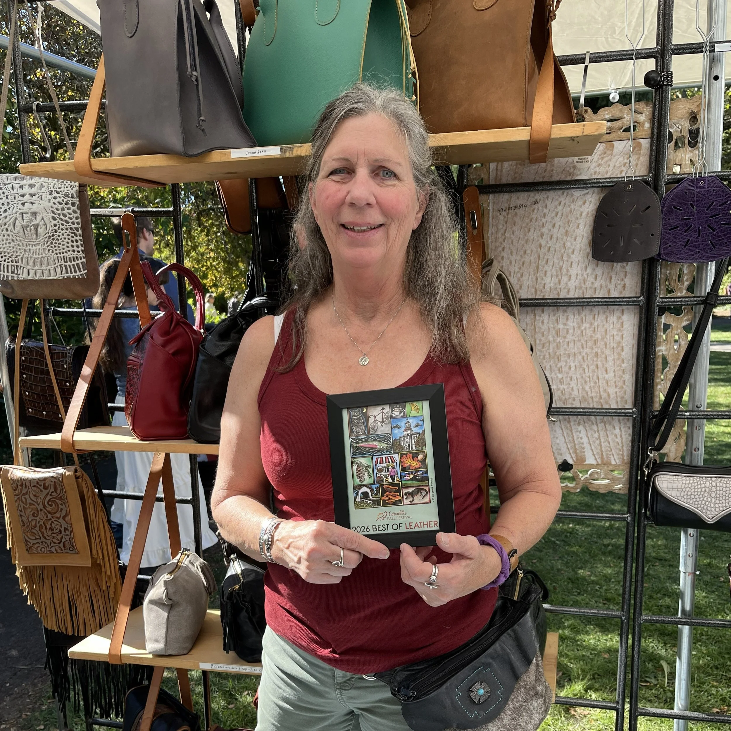 A woman smiling and holding a framed Corvallis Fall Festival poster that reads "2026 best of leather."