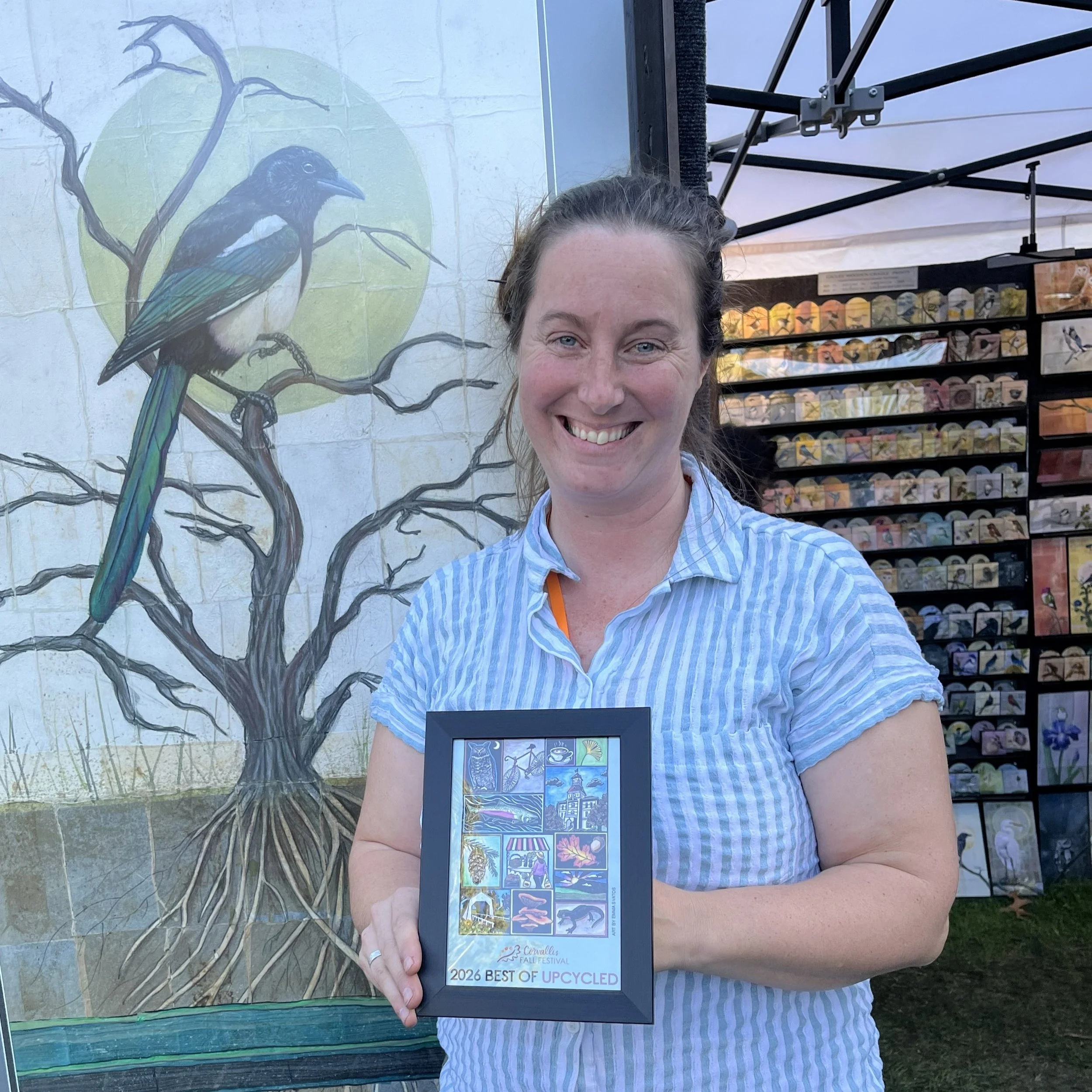 A woman smiling and holding a framed Corvallis Fall Festival poster that reads "2026 best of upcycled."
