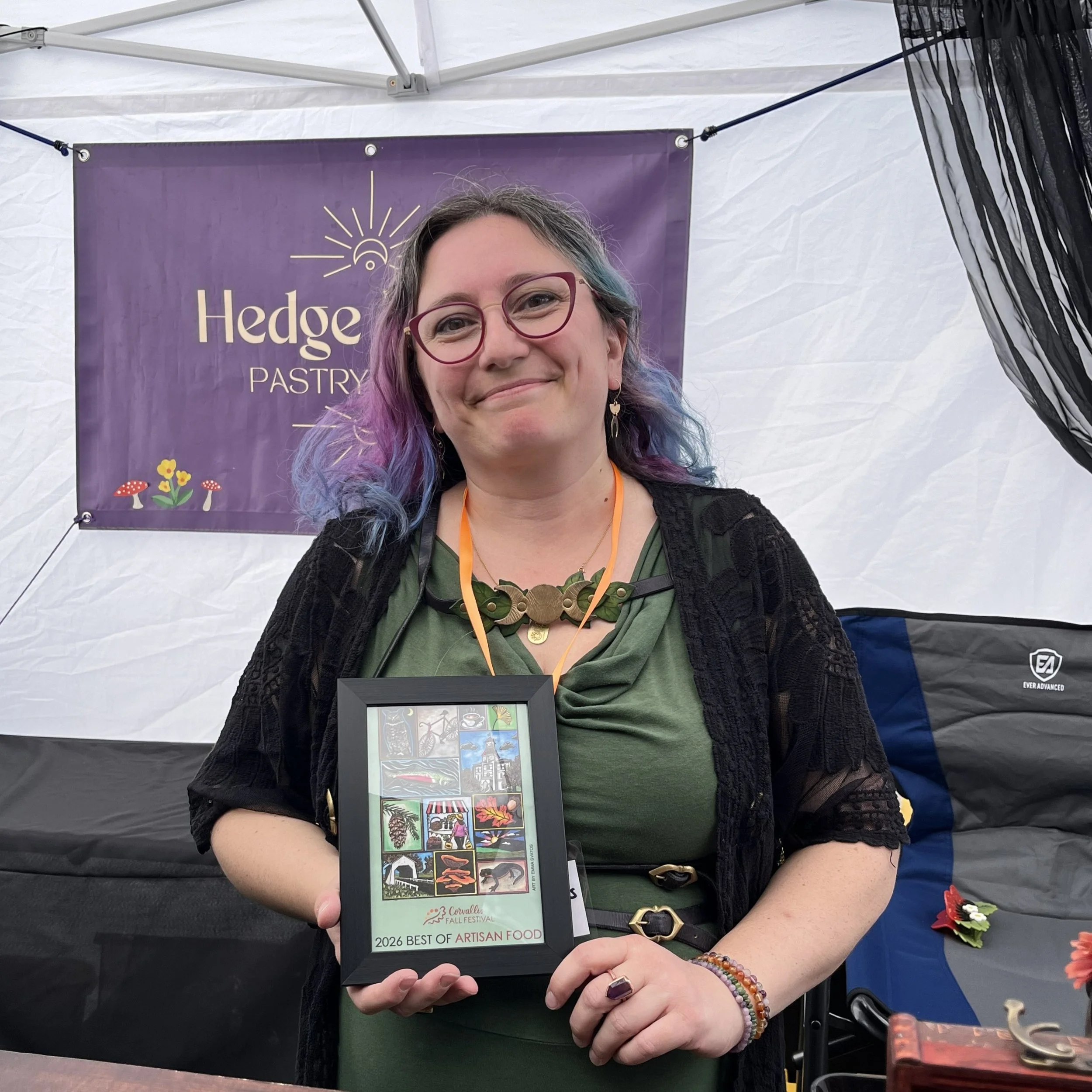 A woman smiling and holding a framed Corvallis Fall Festival poster that reads "2026 best of artisan food."