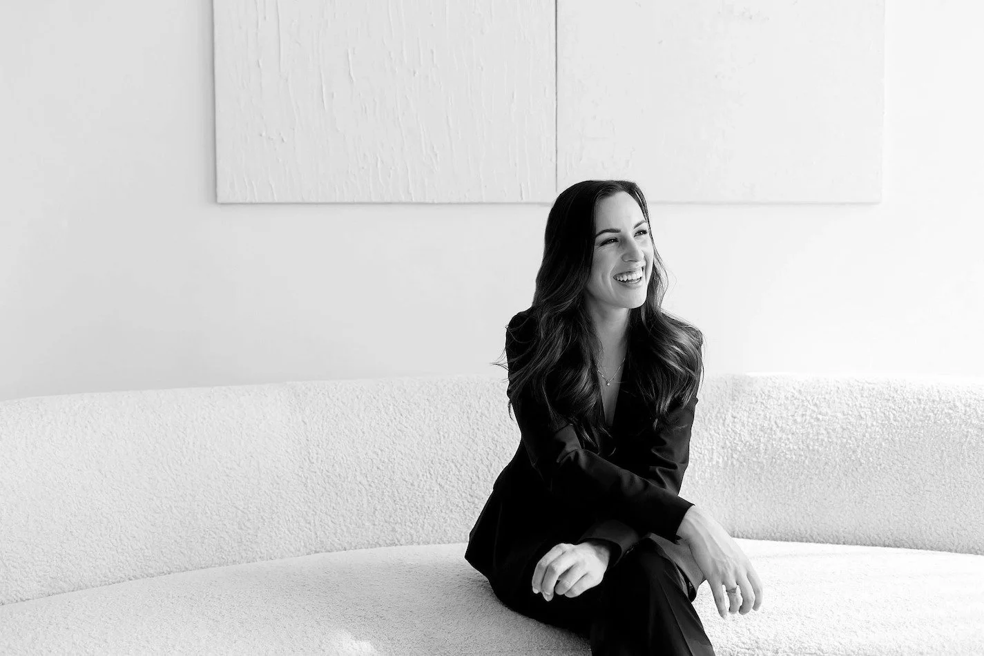 The Modern Ū founder, Rochelle Rudolph-Gonzalez, a woman with long wavy hair sitting on a light-colored textured couch, smiling and looking to the right in a modern, minimalist room.