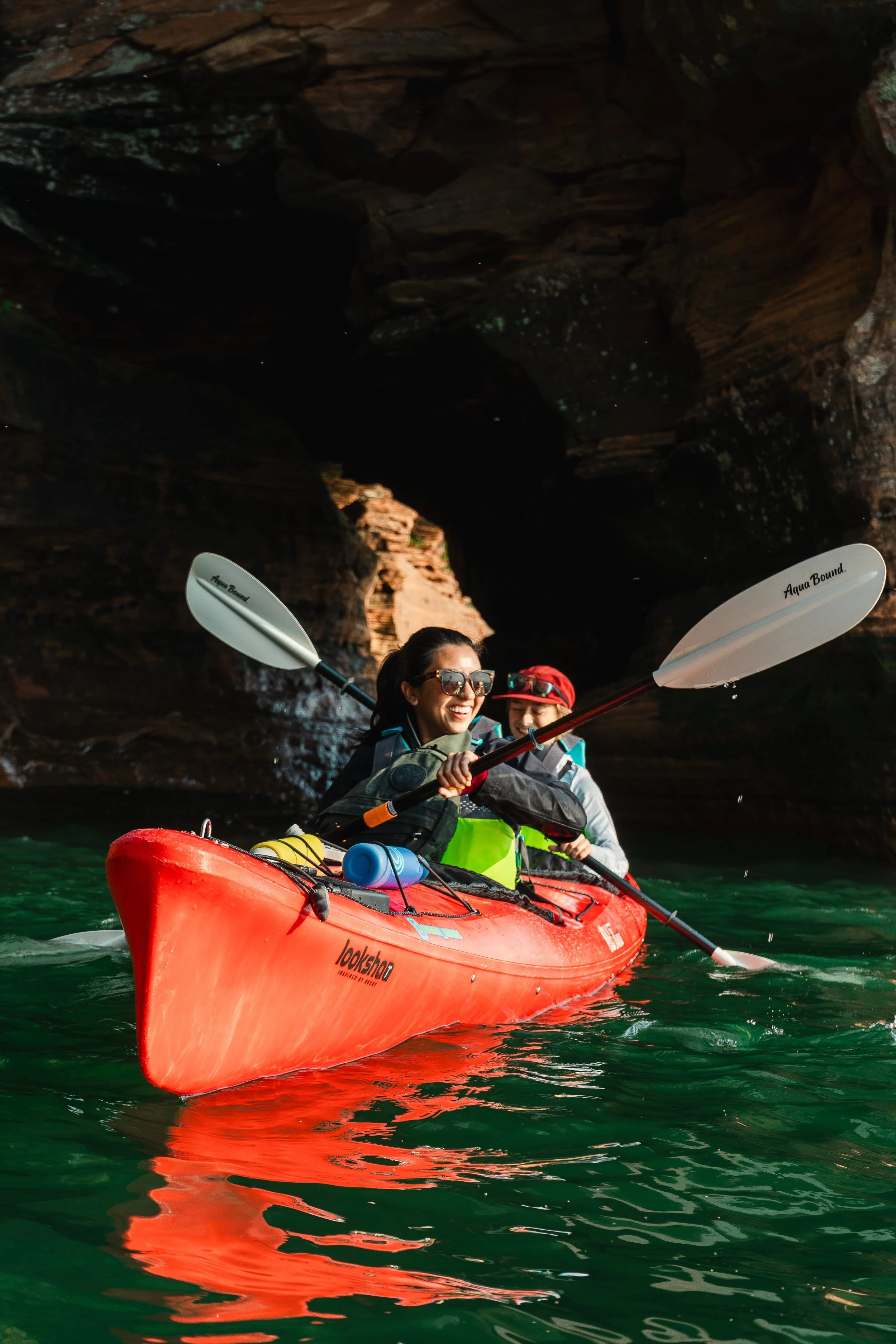 Two people kayaking in a narrow canyon with tall, rocky walls. The person in front is smiling, wearing sunglasses, and holding a paddle. The kayak is red, with water bottles attached to the front.