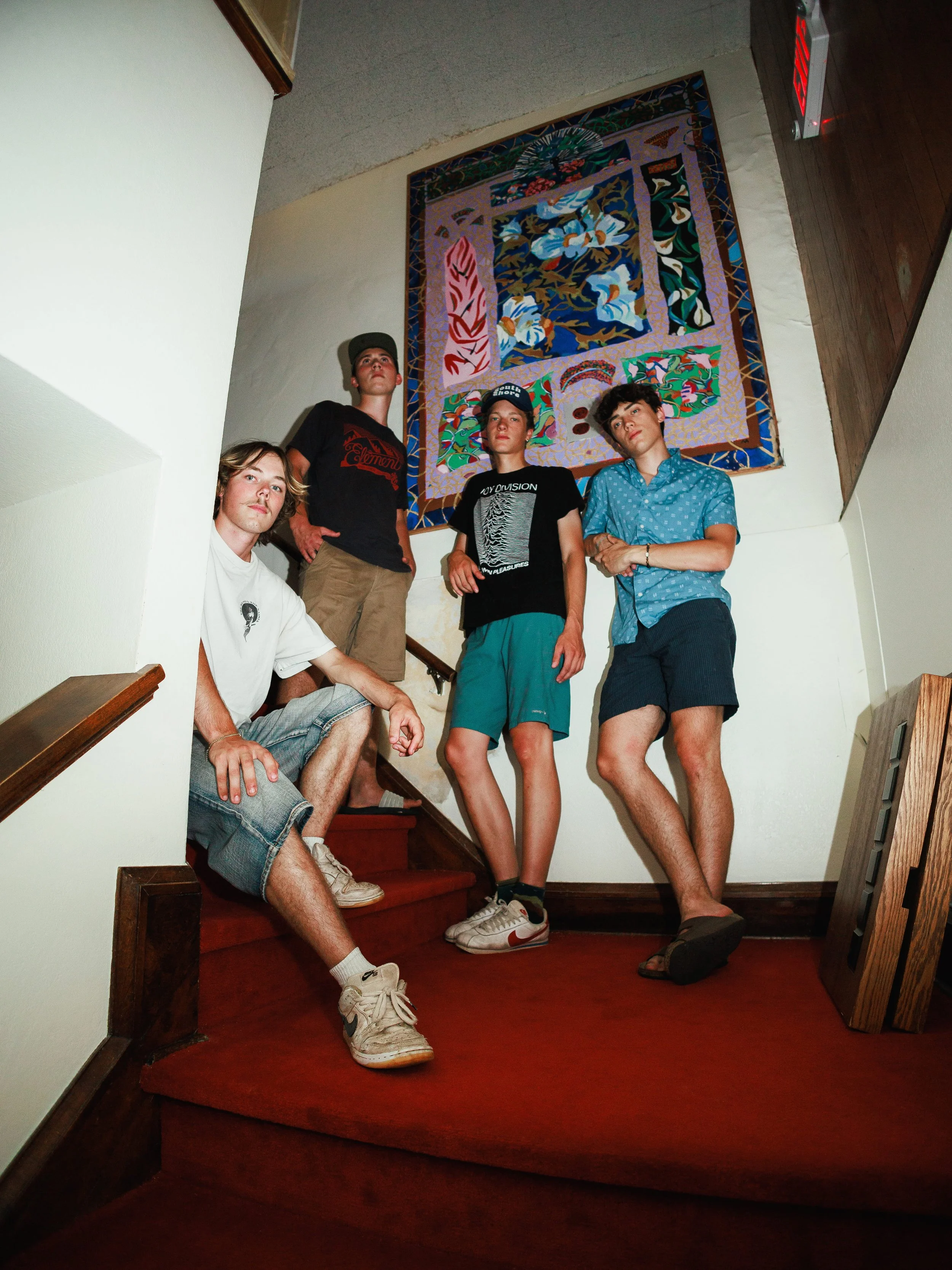 Four young men standing and sitting on a staircase in front of a colorful abstract tapestry. One sitting on the stairs, three standing, dressed in casual summer clothing including t-shirts, shorts, and sneakers.