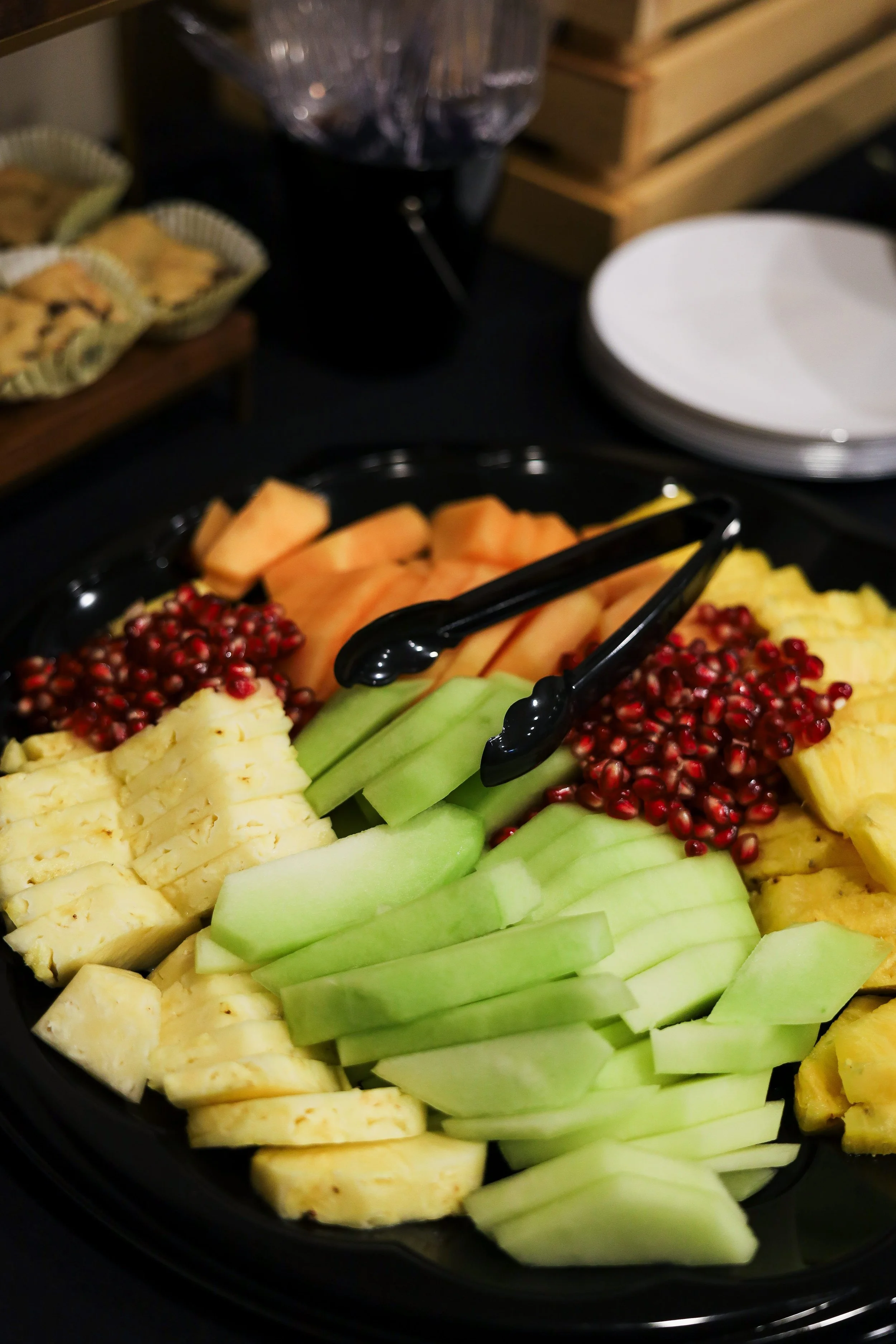 A platter of fresh sliced fruit including pineapple, honeydew melon, cantaloupe, and pomegranate seeds, on a black serving tray with black tongs.