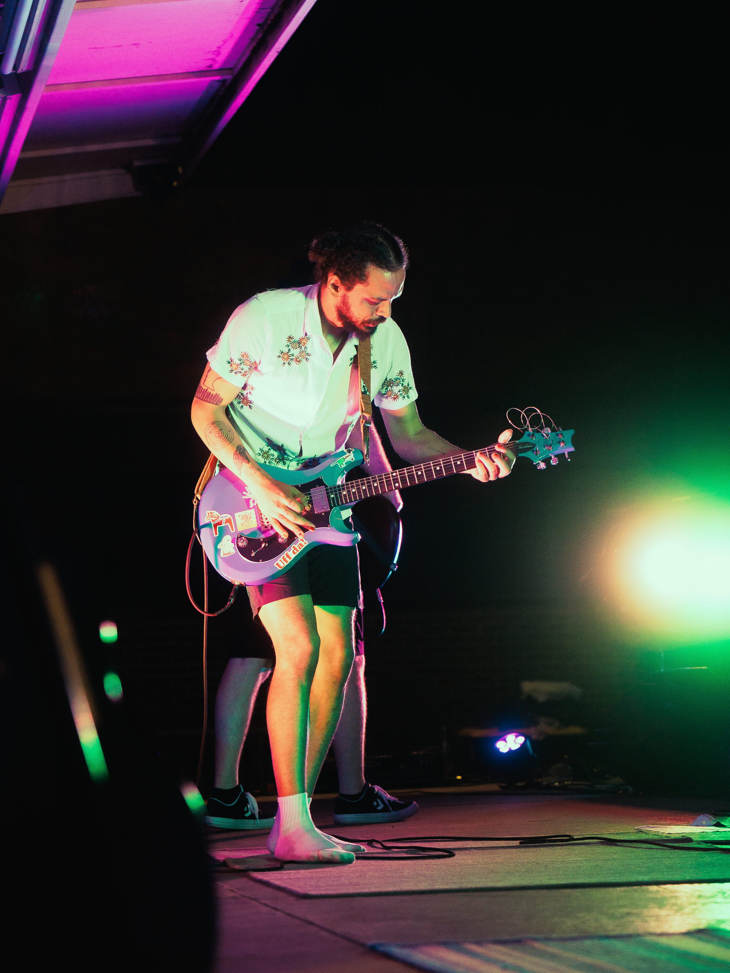 A person with curly hair, beard, and tattoos playing an electric guitar on stage, wearing a floral shirt, shorts, and sneakers, with colorful stage lights illuminating the scene.