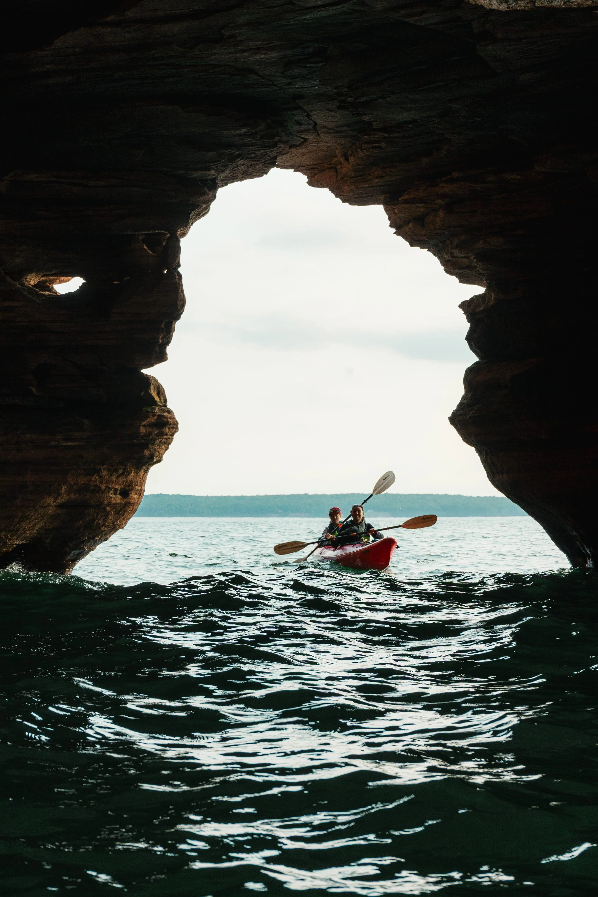 Two people kayaking through a water passage under a rocky overhang with a view of the open water and distant land in the background.