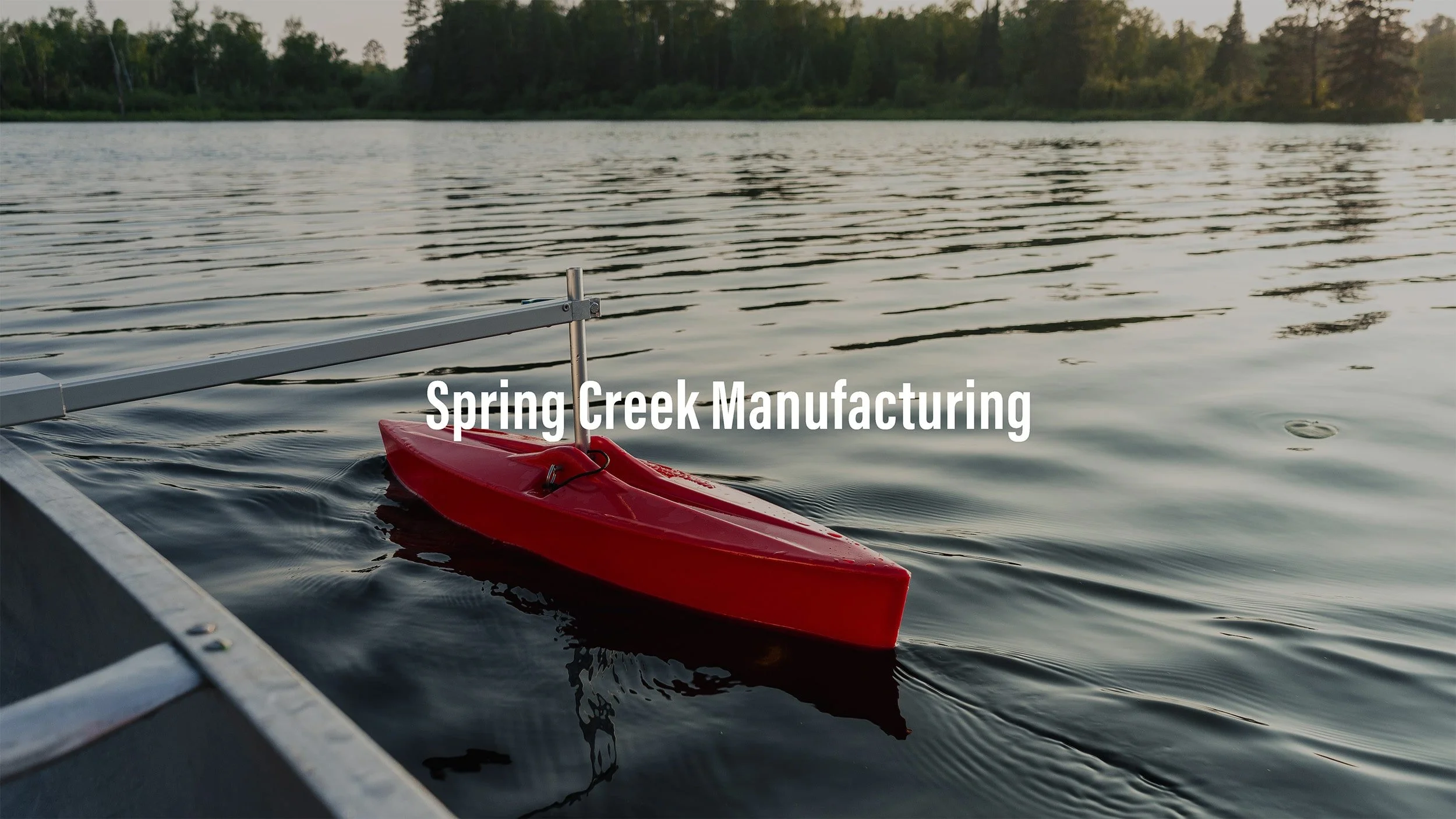 A red model boat attached to a dock on a river, with trees in the background during sunset.