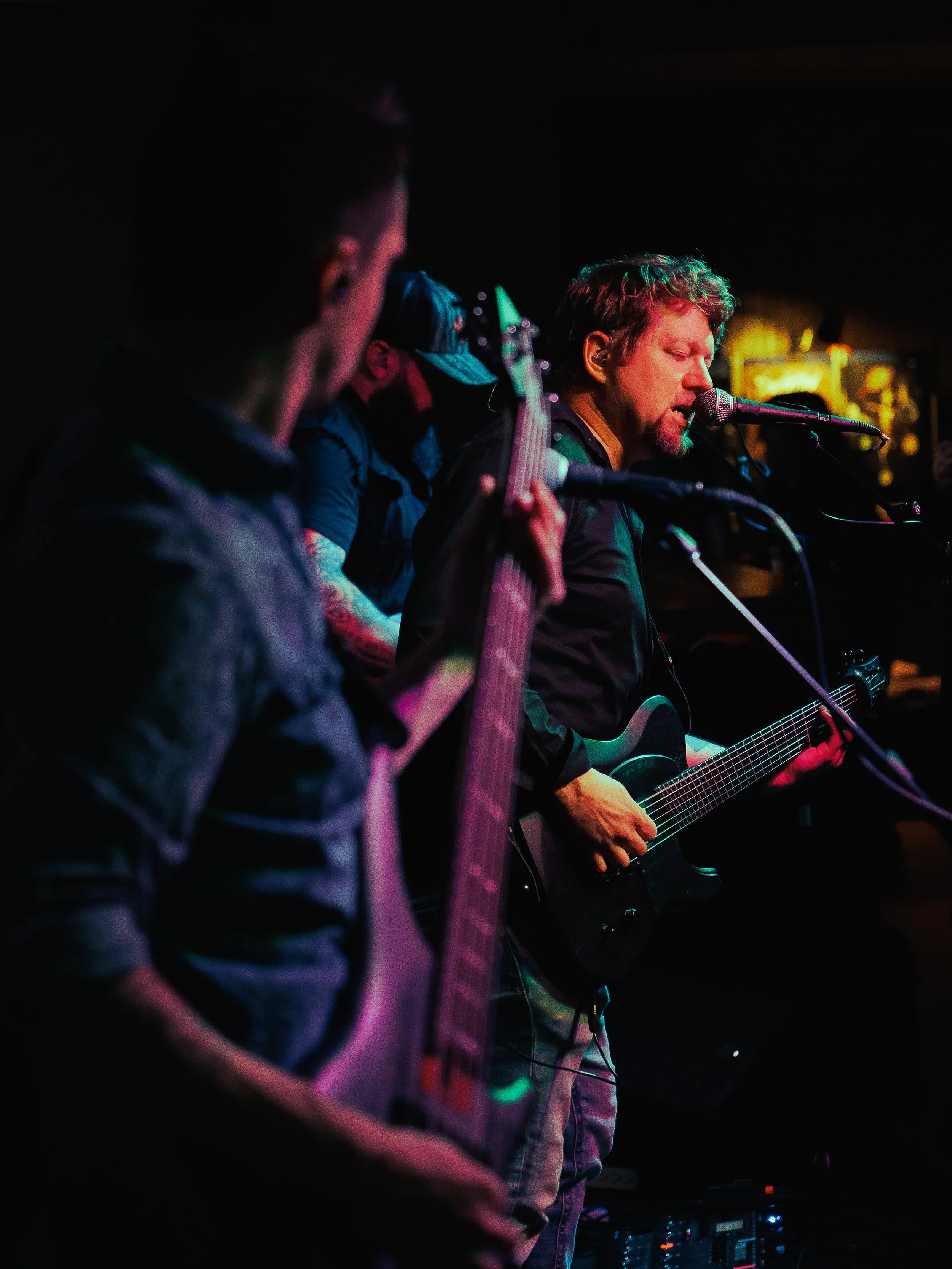 Musicians performing live in a dimly lit bar, with one singer playing an acoustic guitar and another musician playing an electric guitar.