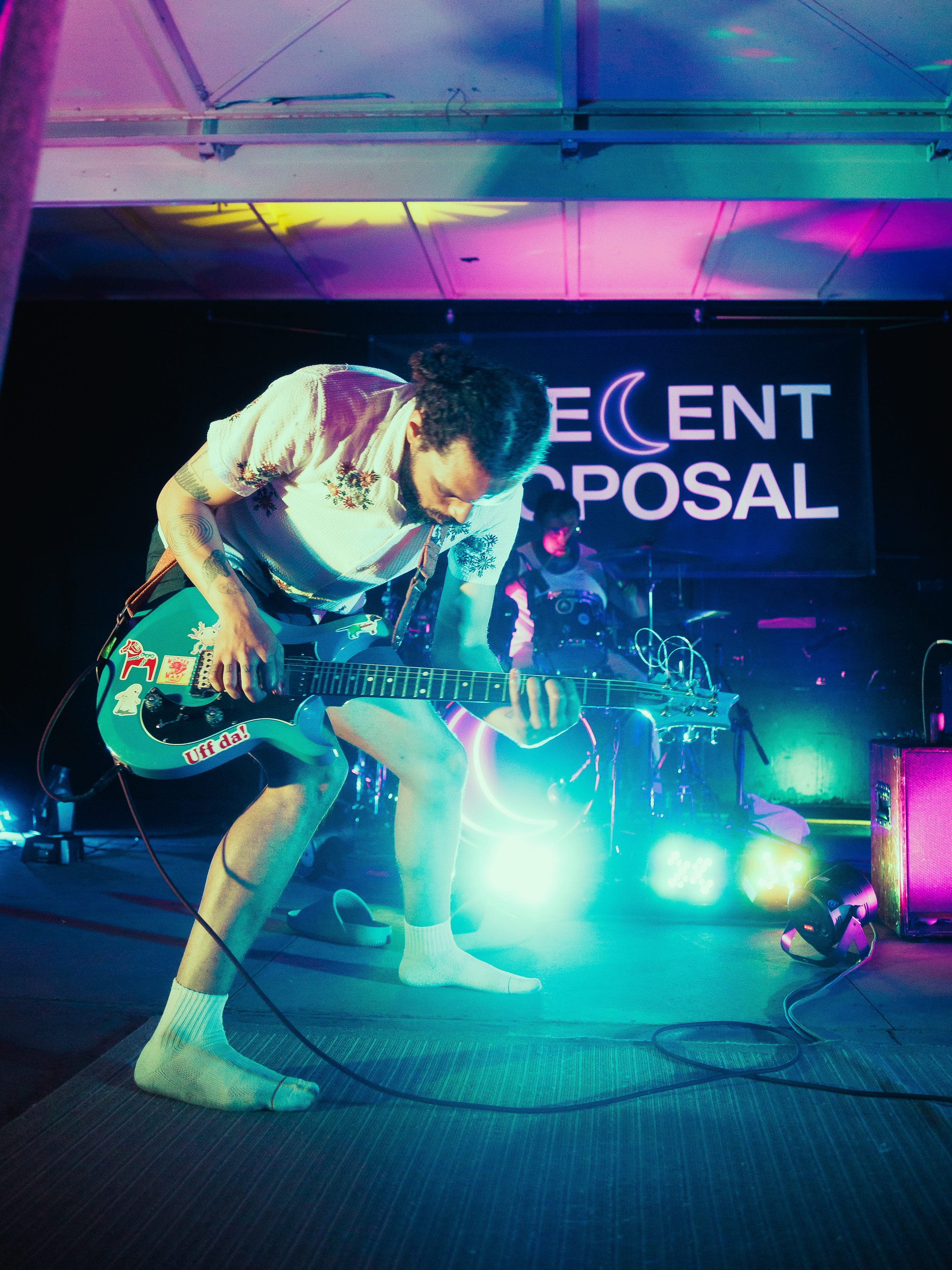 Musician playing guitar on stage with colorful lights and a sign that says 'Recent Proposal' in the background.
