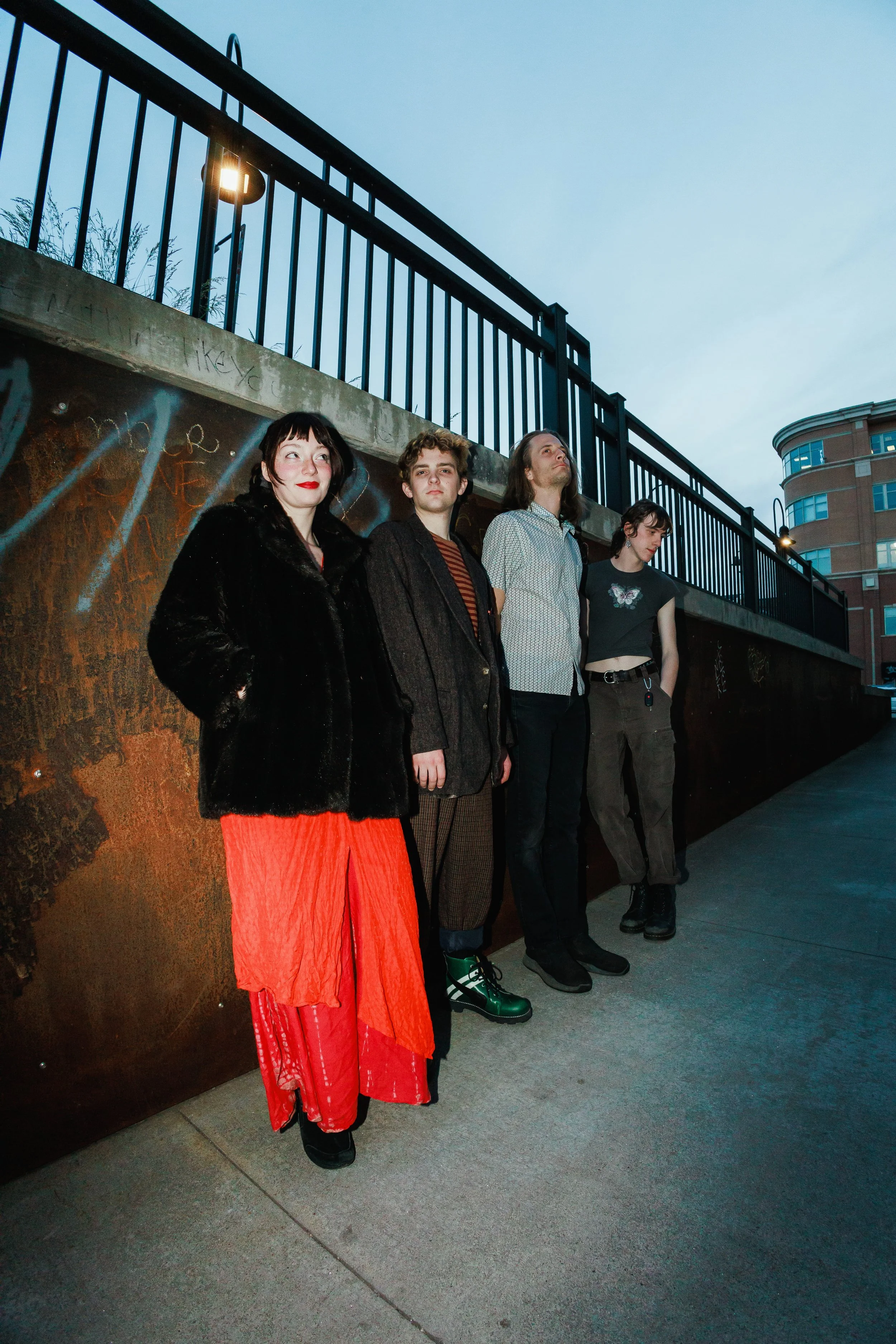 Four young adults standing against a graffiti-covered wall on an urban street during the evening, with a clouded sky and city buildings in the background.