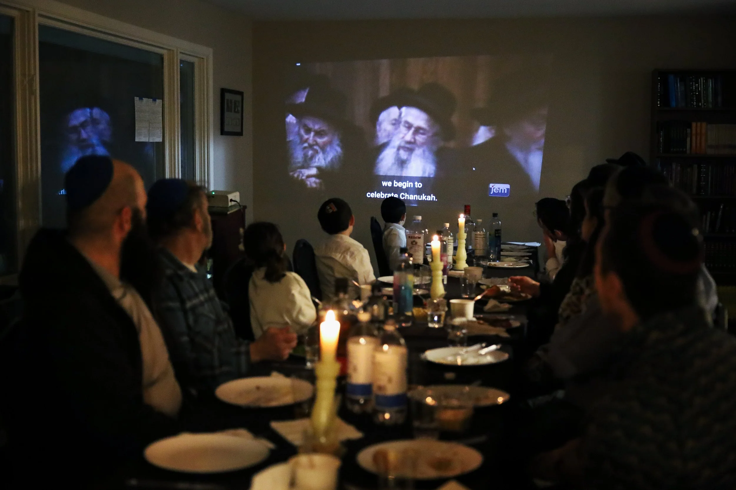People gathered around a table watching a projected image on the wall that shows elderly men with beards and black hats, with a caption that reads 'we begin to celebrate Chanukah.' The table has candles, bottles, and dishes in a dimly lit room.