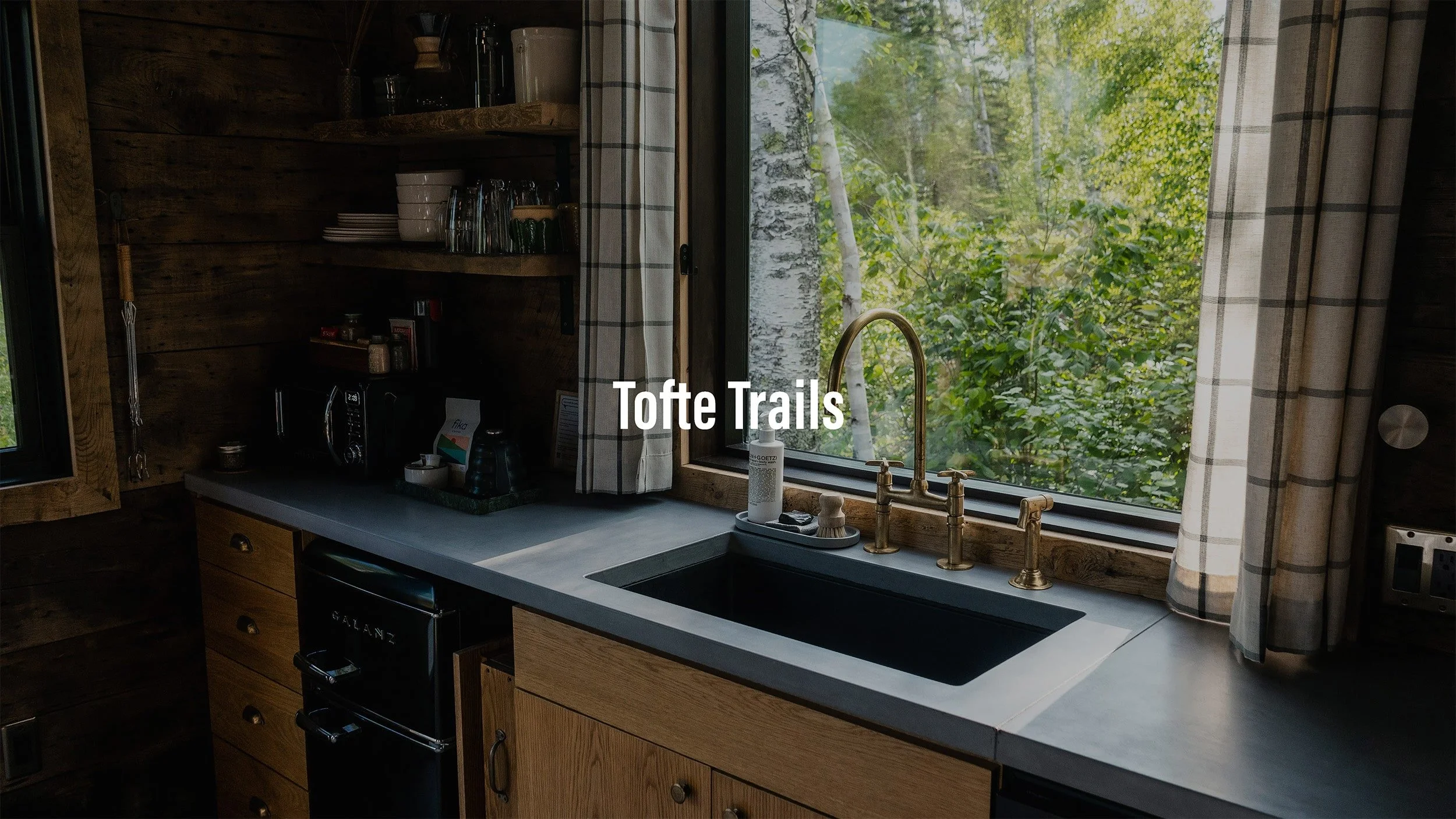Kitchen with a window showing green trees outside, gold faucet, black sink, wooden cabinets, and shelves with dishes and glassware.