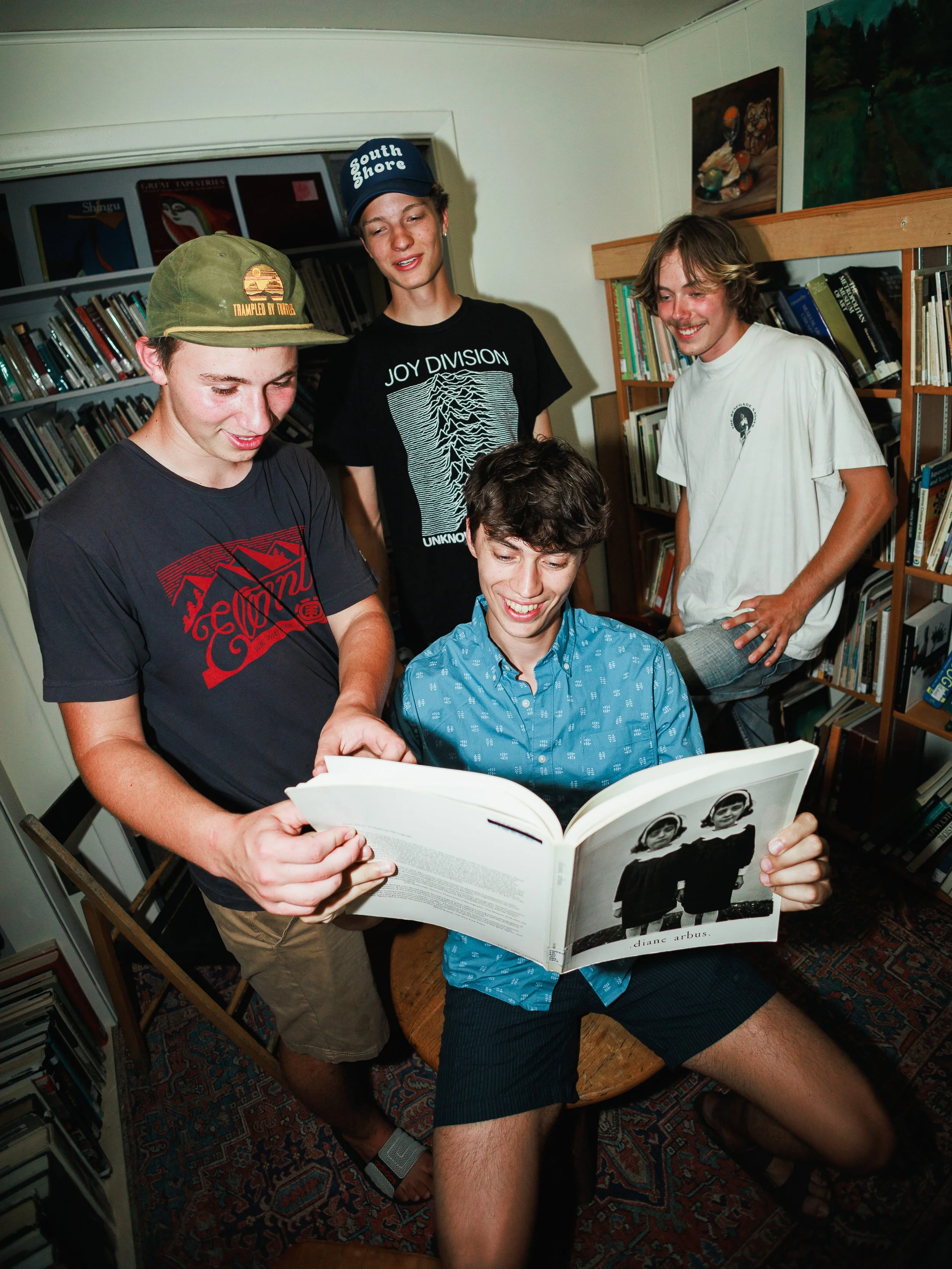 Four young men in a bookstore, one sitting on a wooden stool reading a book with black and white photos, the others standing around him, smiling and looking at the book.