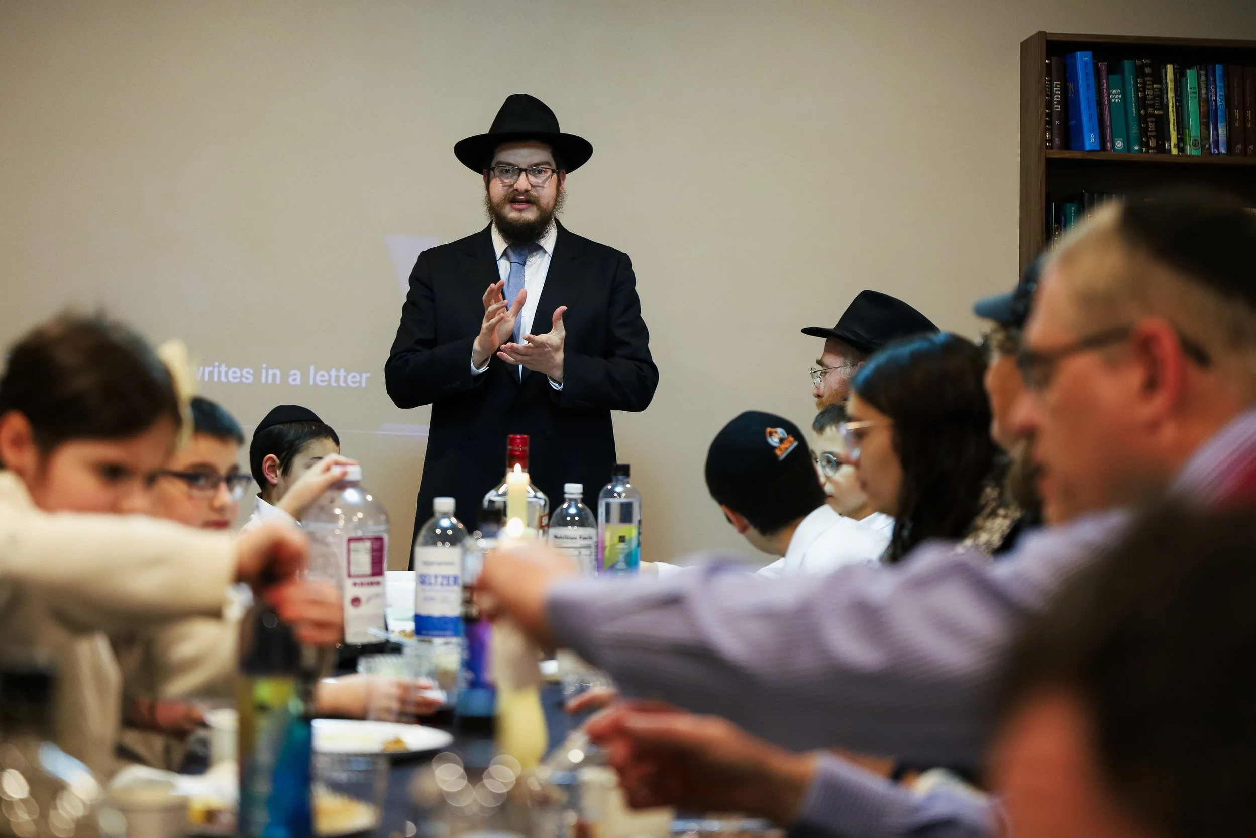 Man dressed as a rabbi with a black hat and suit giving a talk to a group of children and adults seated at a table with bottles and food.