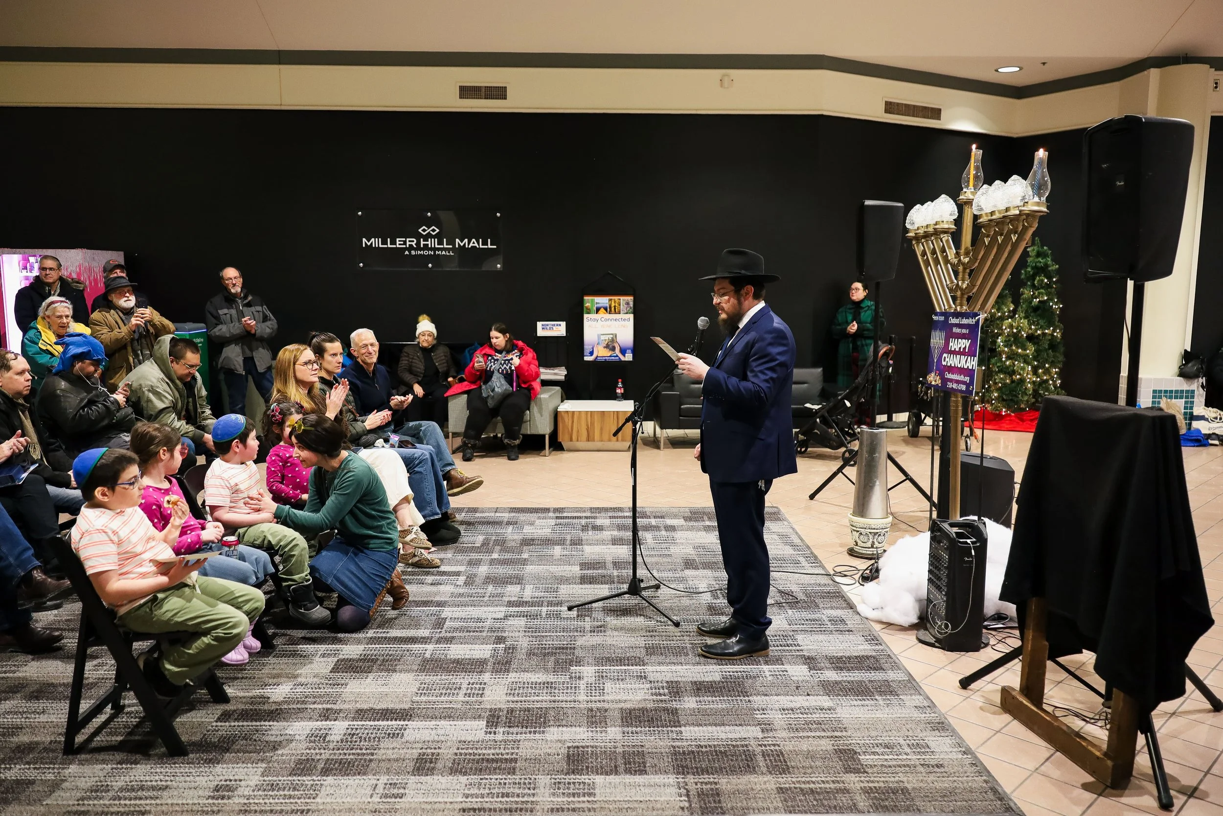 A man dressed in a dark suit, hat, and glasses speaking into a microphone at a gathering in a mall, with children and adults seated and standing around him, decorated for Hanukkah and Chanukkah celebration signs visible.