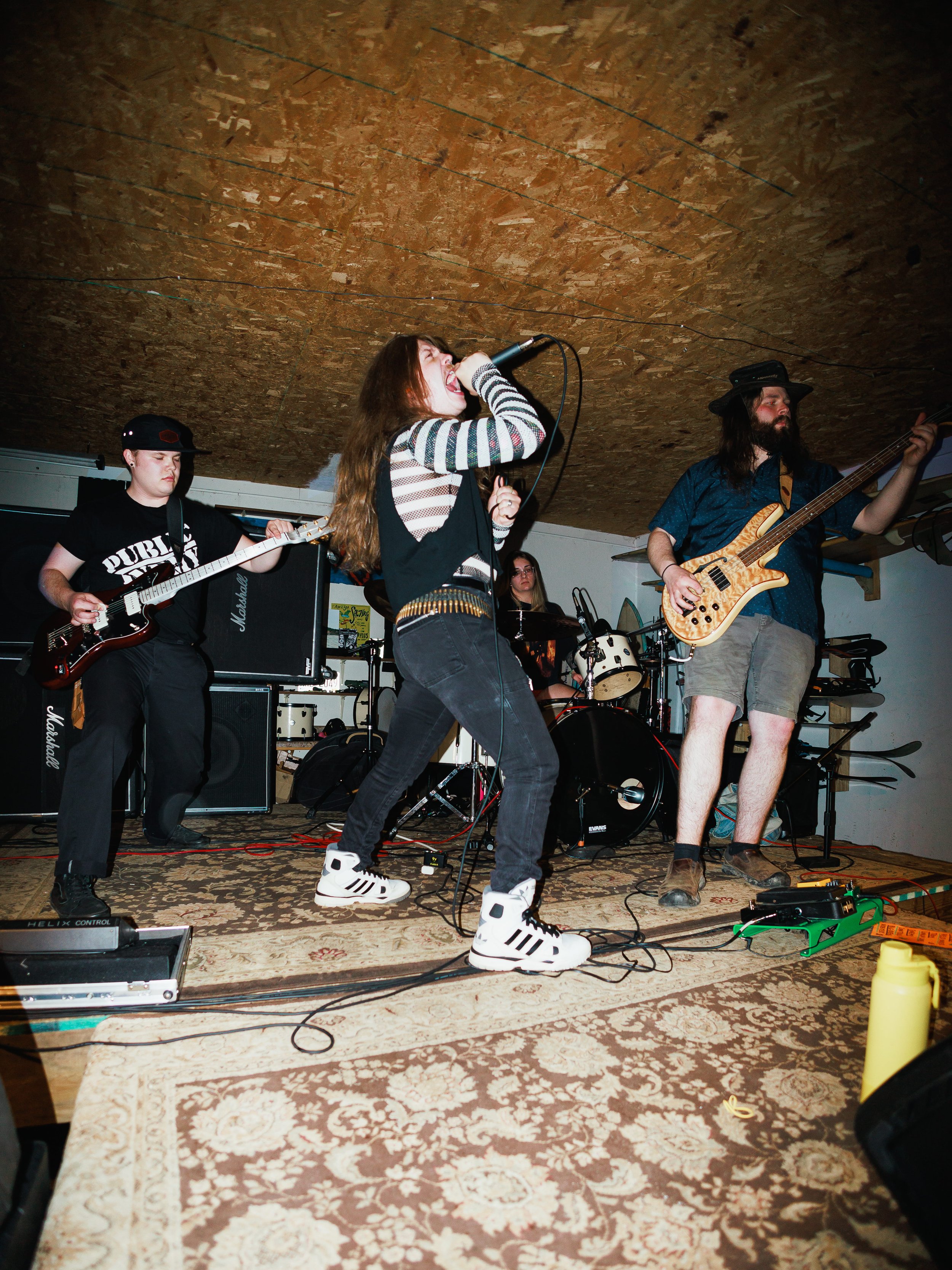 A band performs on stage with a female singer in the center, two guitarists on either side, a drummer in the background, and music equipment including amplifiers, pedals, and microphones.